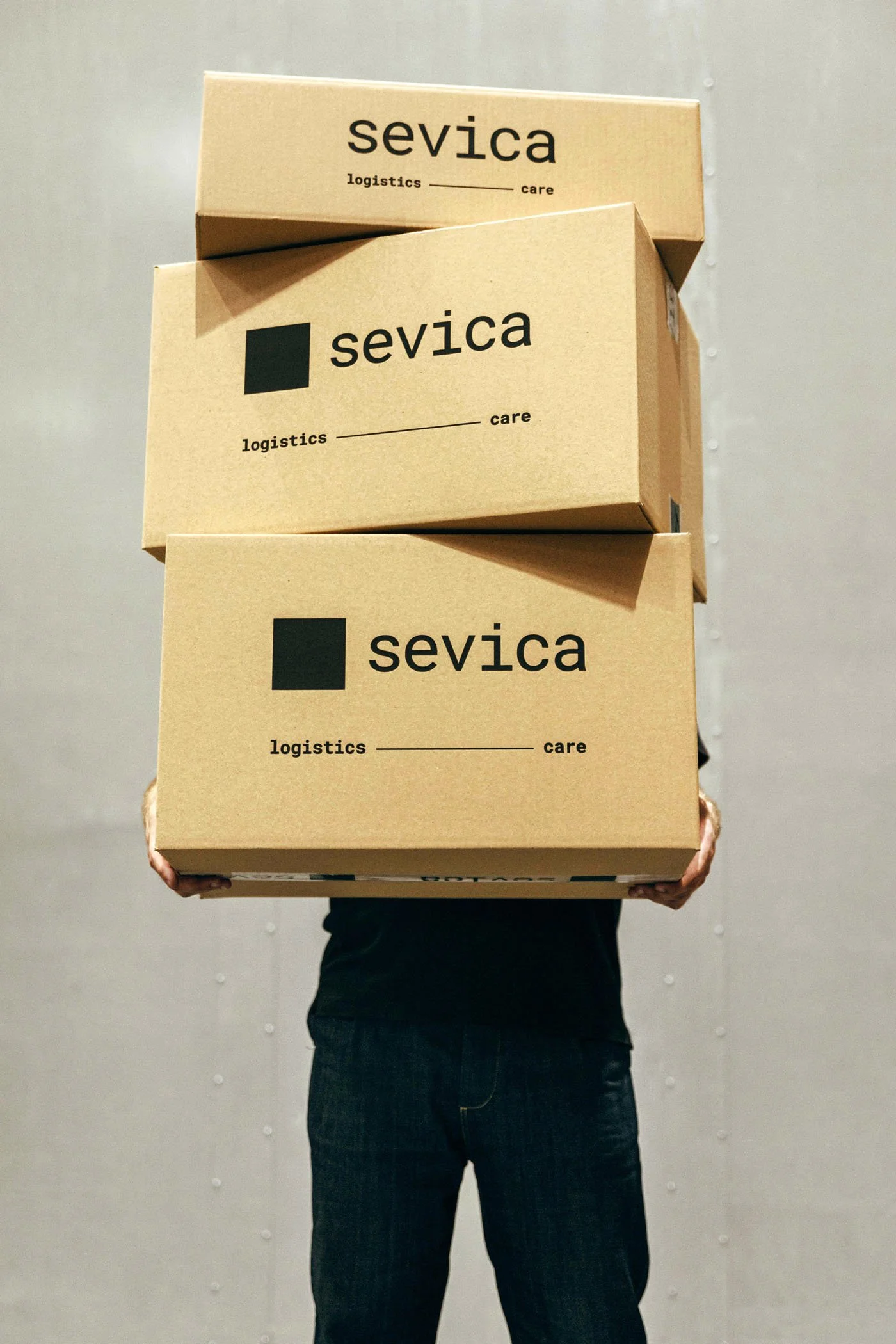 Persona sosteniendo varias cajas de cartón con la marca 'sevica' y el texto 'logistics care', en un entorno interior.