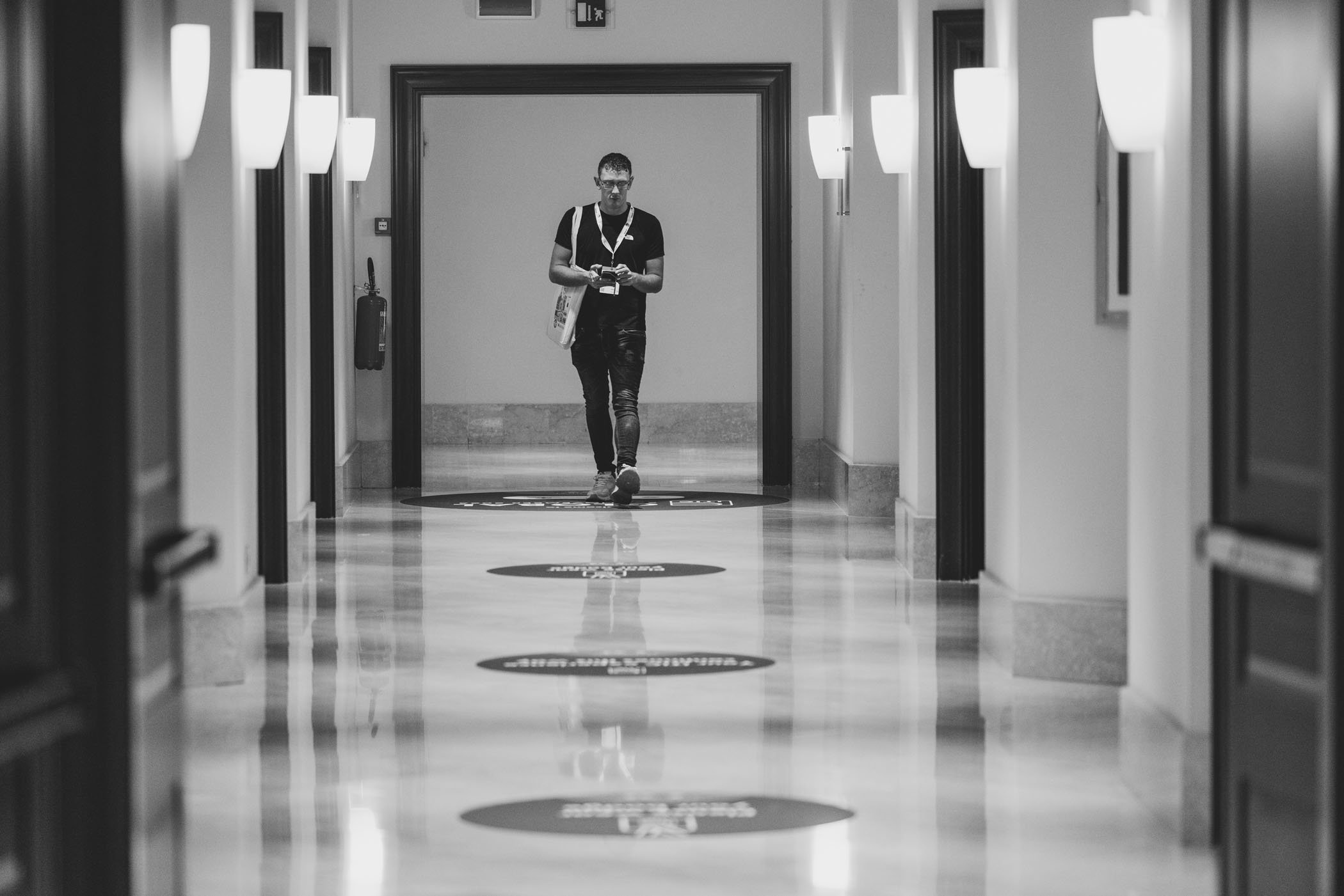 Persona caminando en pasillo de hotel en blanco y negro, con puertas a los lados y luces en las paredes.
