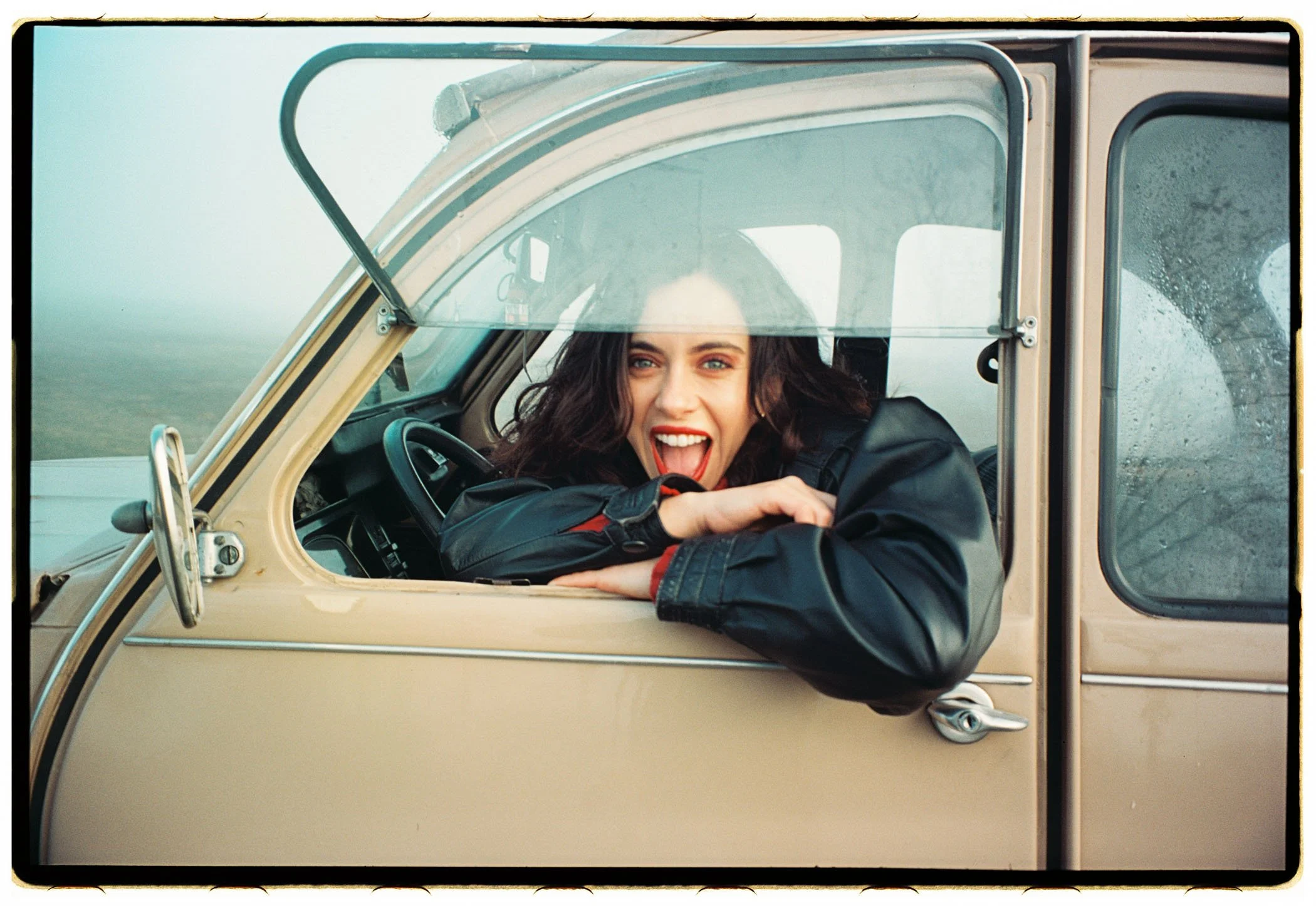Mujer sonriente con cabello oscuro a la windsor, con chaqueta negra, asomada por la ventana de un vehículo vintage beige, en un paisaje de campo con cielo nublado.