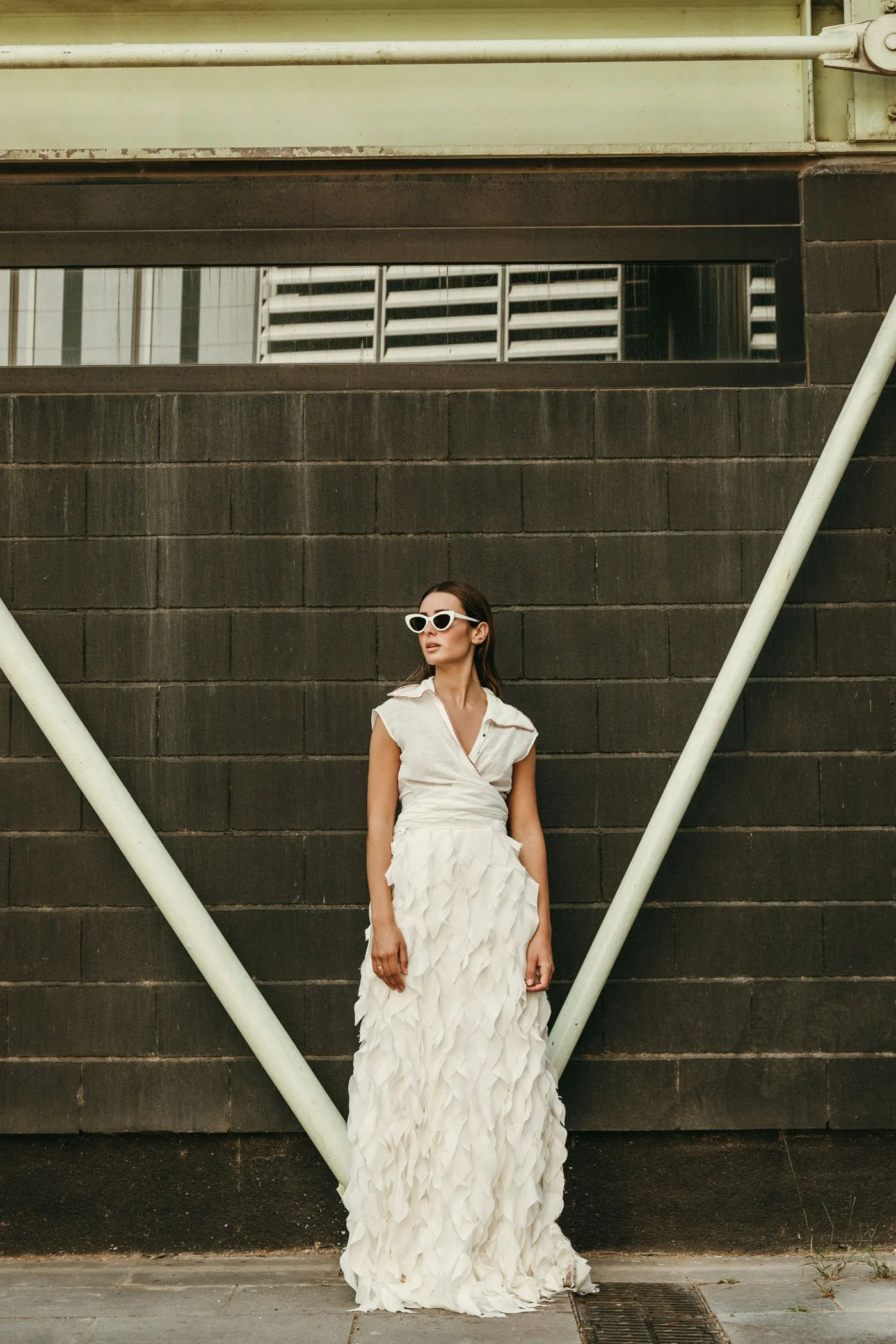 Mujer con vestido blanco y gafas de sol de pie contra una pared de bloques oscuros.