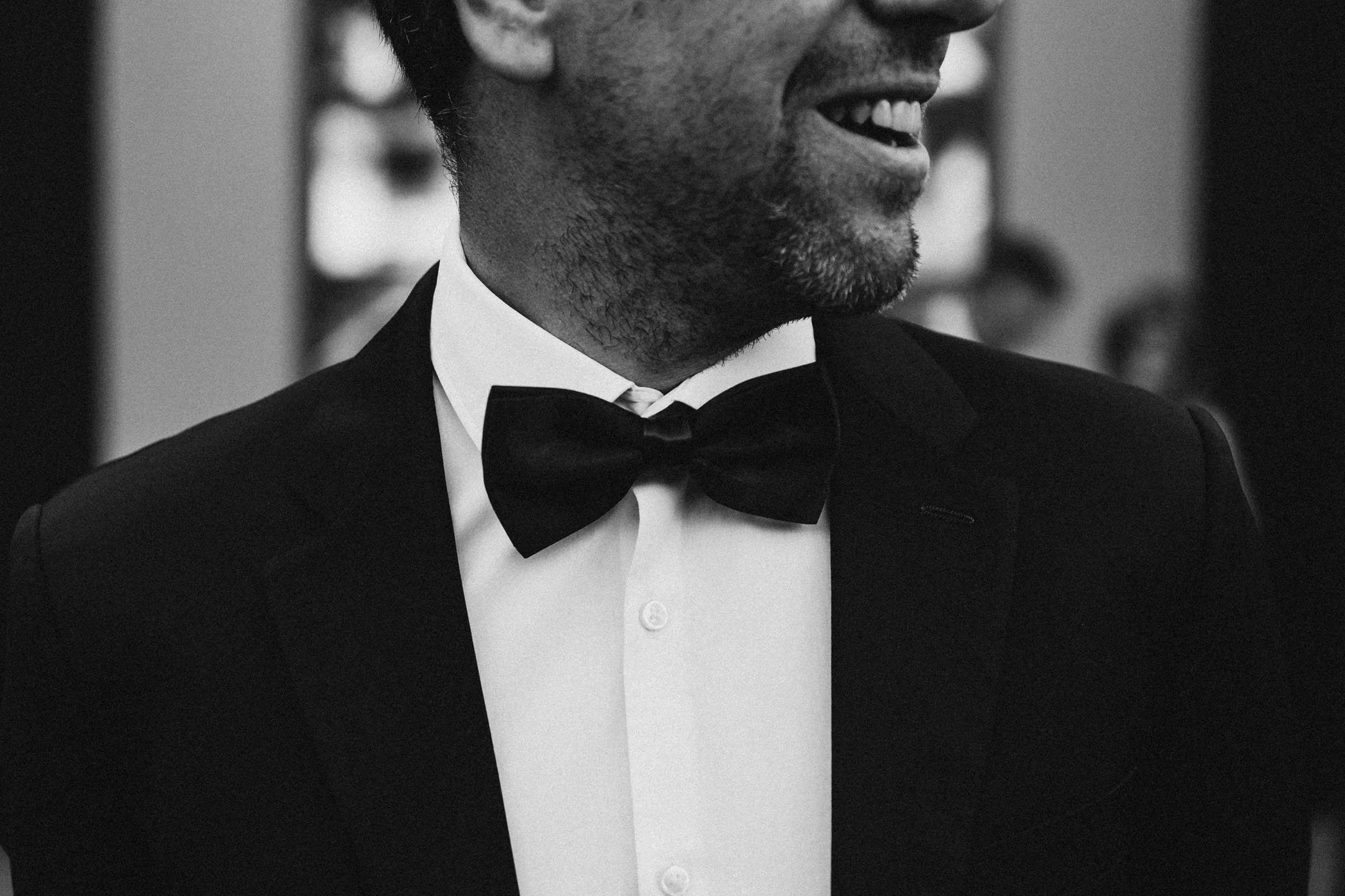 Hombre con barba vestido con esmoquin y pajarita, sonriendo, en un entorno formal en blanco y negro.