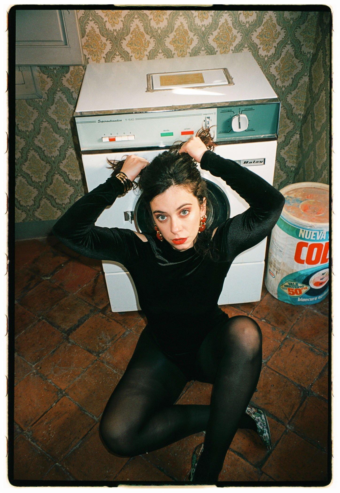 Una mujer joven con ropa negra, pantalones de malla negra, tacones peep toe con estampado y aretes grandes rojos, está sentada en el suelo de un piso de ladrillo, con las piernas cruzadas. Ella tiene el cabello oscuro, rizado y lleva maquillaje llama