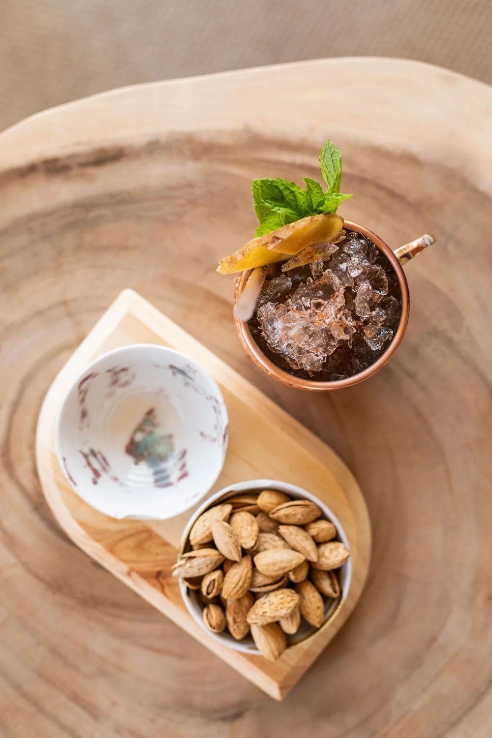 Imagen de una bebida en un vaso con hielo, decorada con una hoja de menta y una rodaja de limón, acompañada de un cuenco con pistachos y una taza vacía, todo sobre una bandeja de madera sobre una mesa de madera.