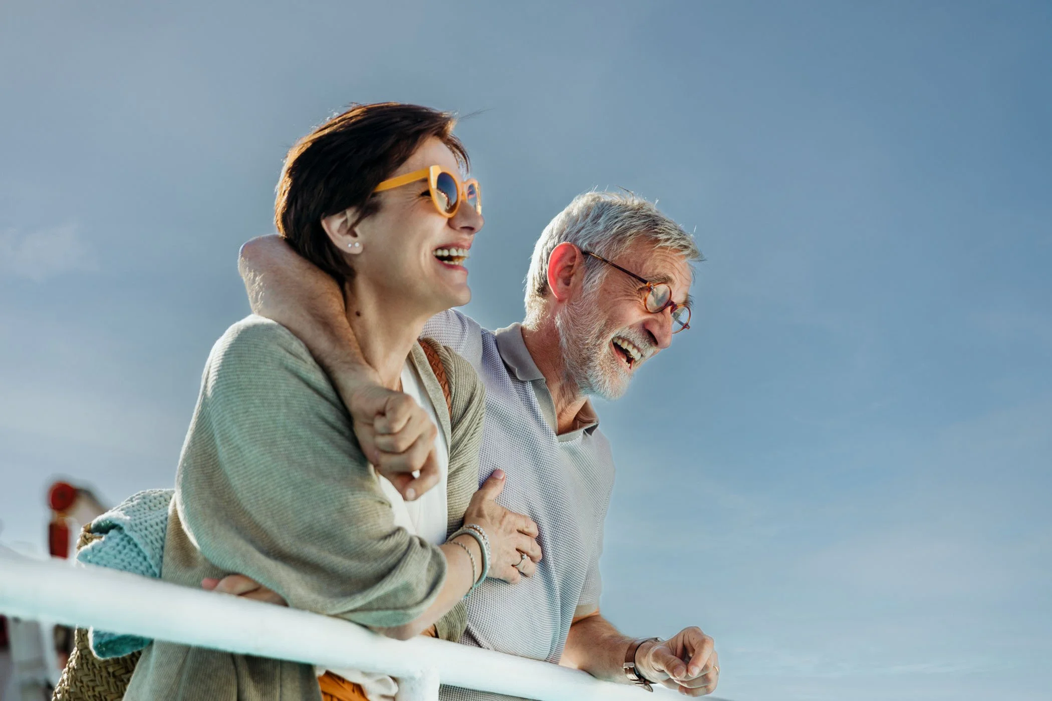 Una mujer y un hombre mayores riendo y disfrutando juntos en un día soleado, apoyados en la barandilla de un barco.