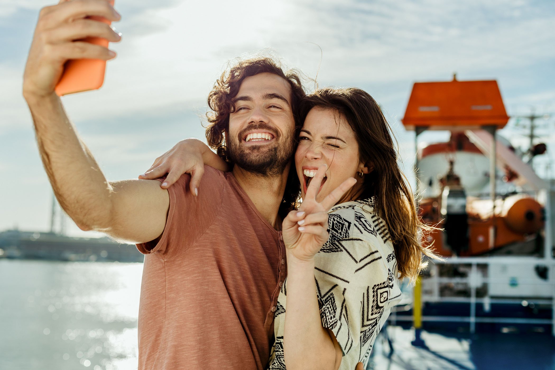 Una pareja joven se toma una selfie con un muelle y un barco en el fondo, ambos sonrientes y felices, haciendo una señal de paz.