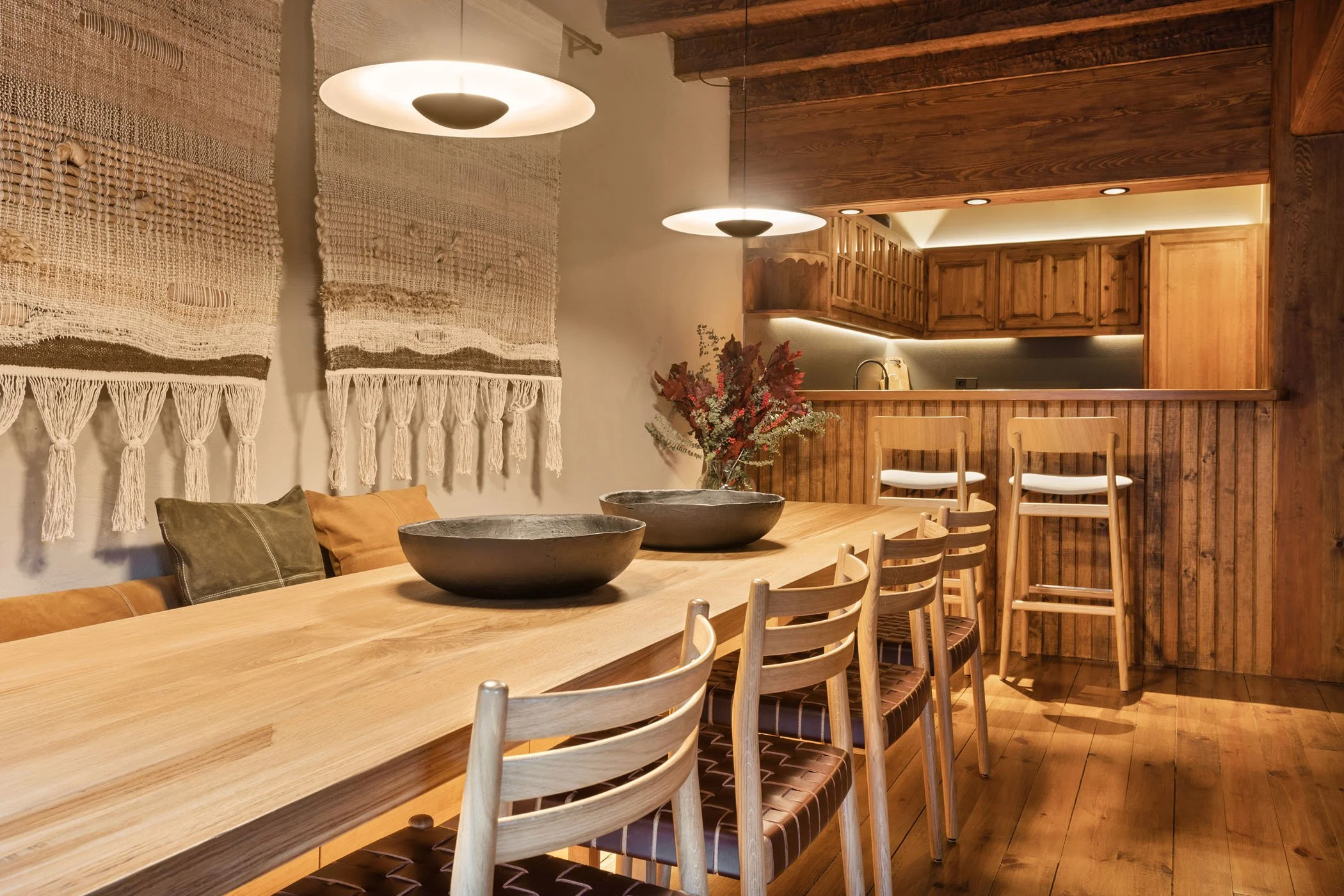 Sala de comedor con mesa de madera, sillas de madera con cojines, decoración en tonos cálidos, cortinas de hilo y lampara colgante en un espacio con diseño rústico.