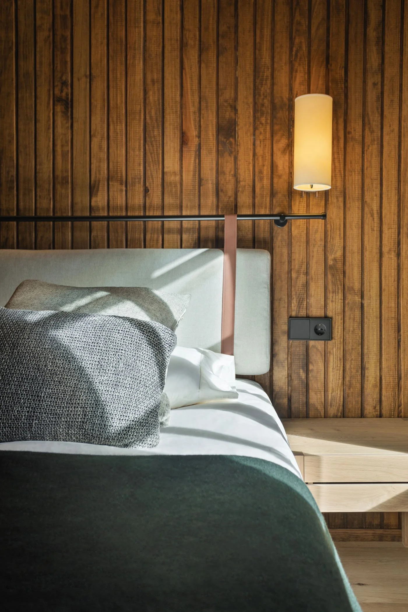 Cama con cabecera acolchada junto a pared de madera, lámpara de pared, enchufe y mesita de madera