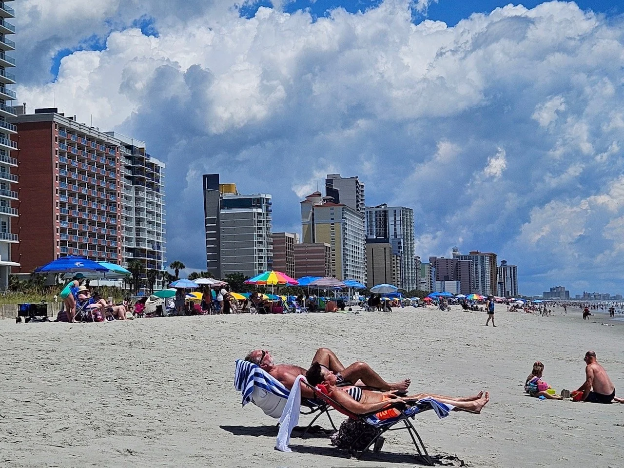People relaxing on the beach with umbrellas and lounge chairs, high-rise buildings in the background, partly cloudy sky.