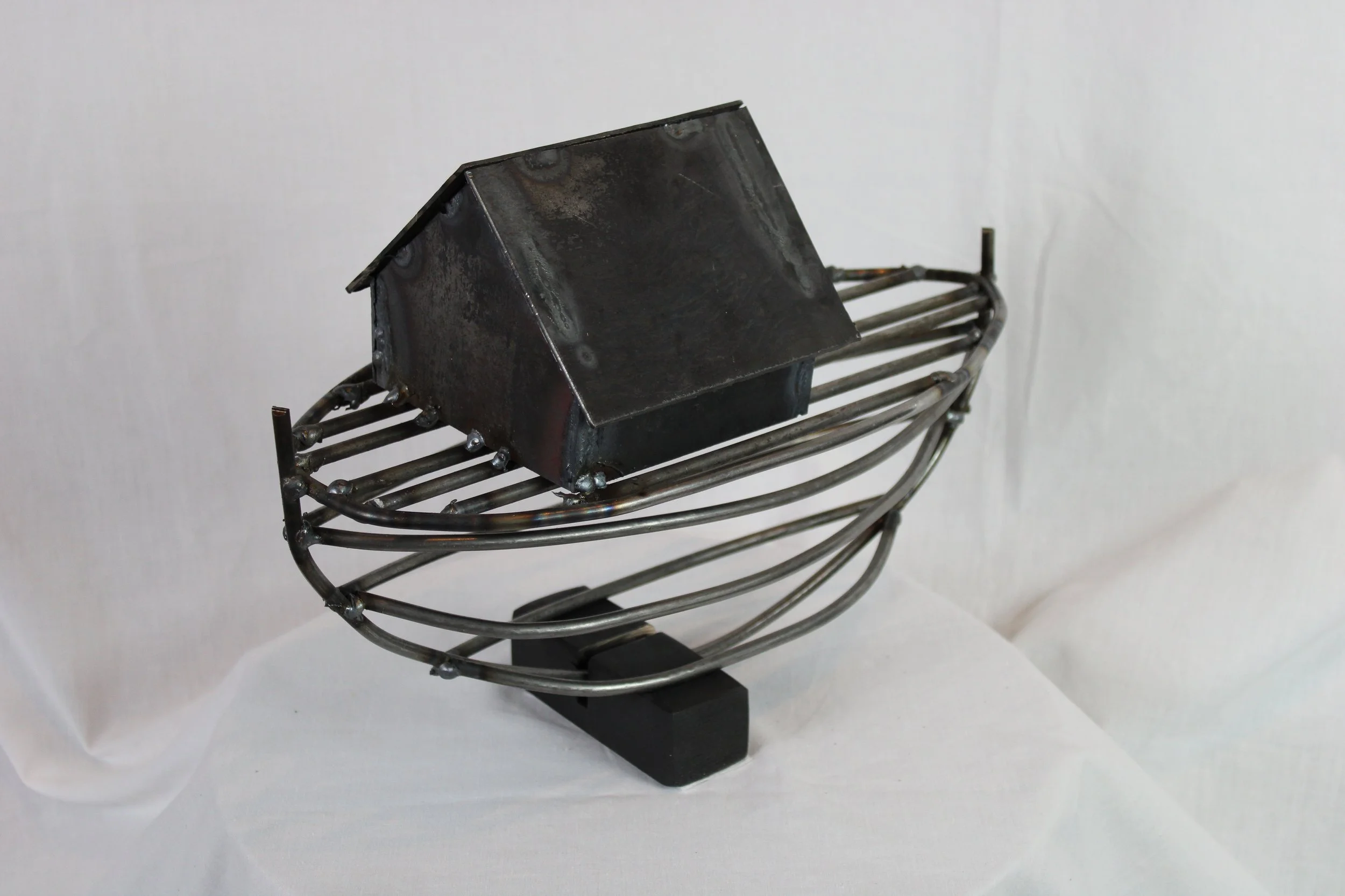 A metal sculpture resembling a boat with a small metal house on top, made from metal rods and sheets, mounted on a black rectangular base.