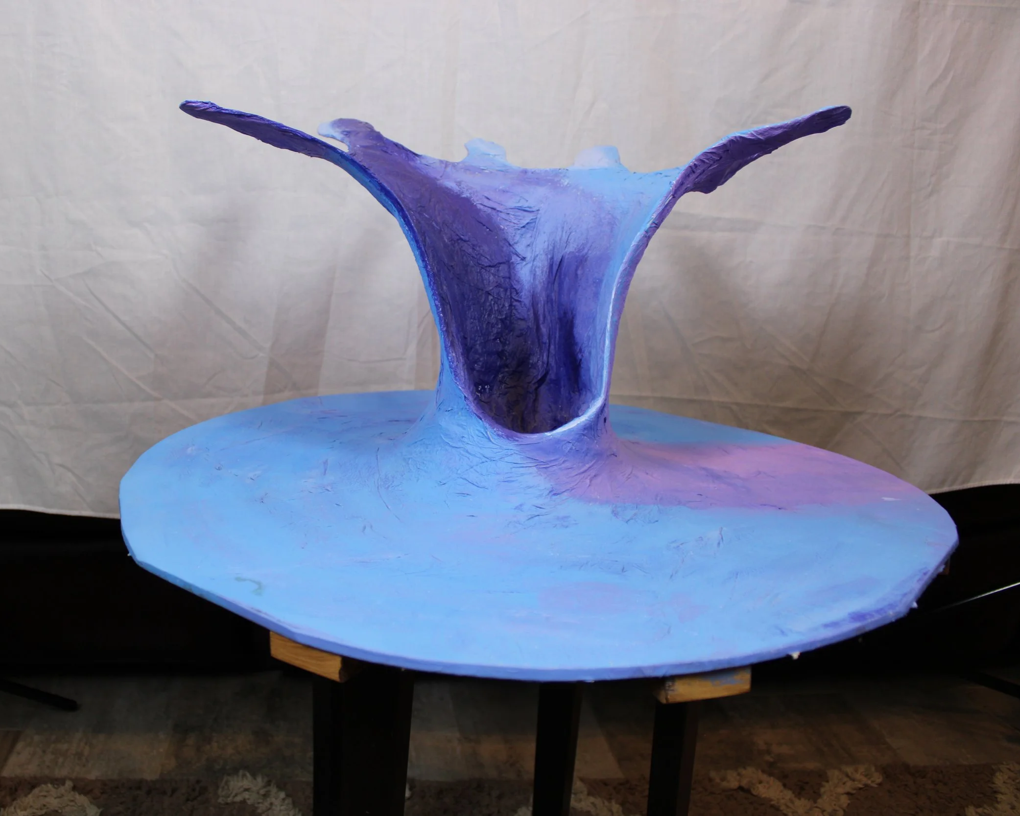 A sculptural piece resembling a whale's tail emerging from the ocean, painted in shades of blue, purple, and pink, placed on a wooden table.