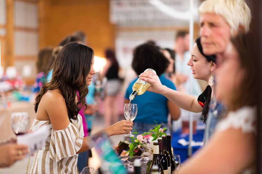 A woman at a wine tasting event pours white wine into a glass for a young woman with long dark hair.