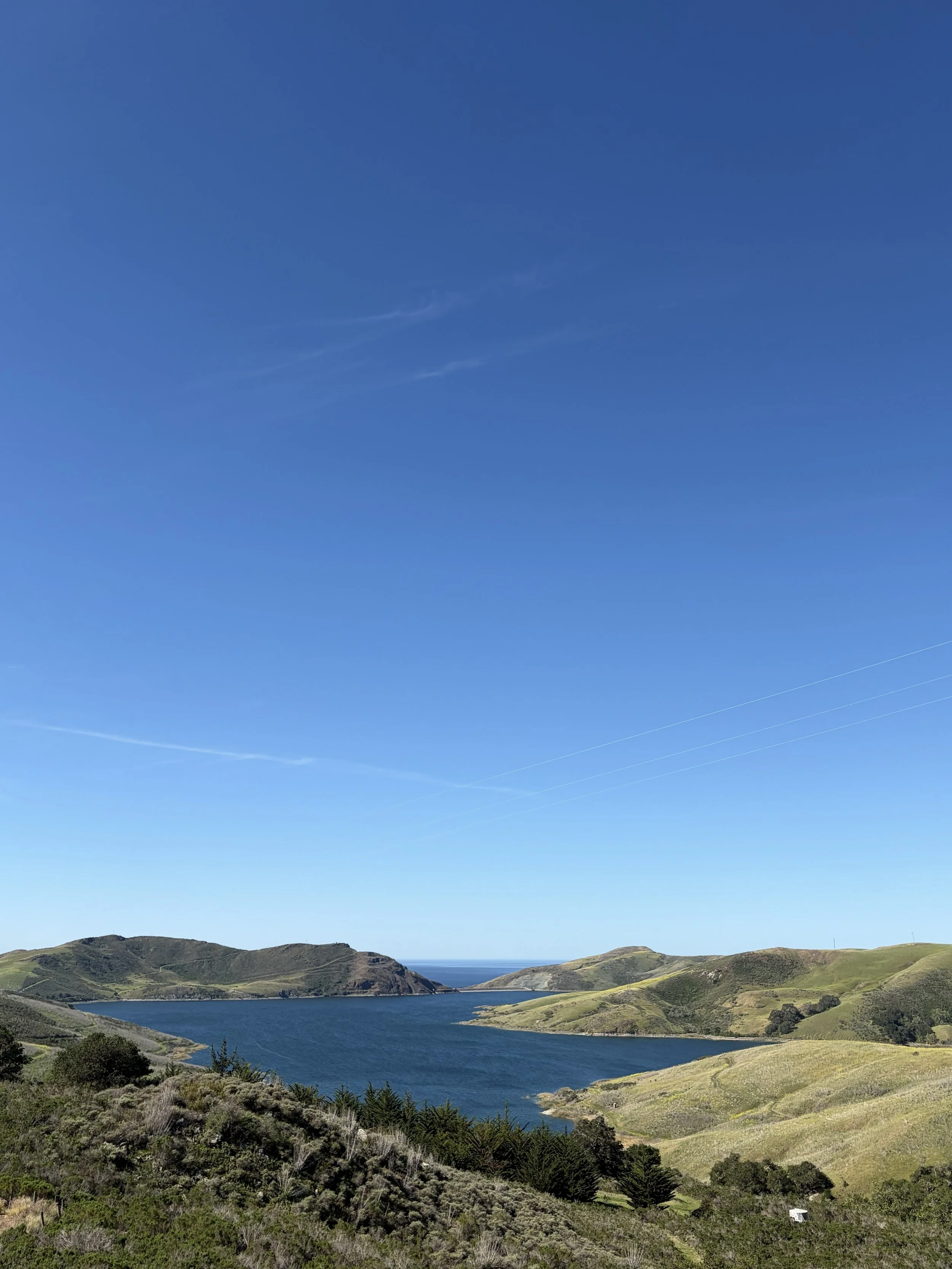 Scenic landscape with a wide water body, rolling green hills, and a bright blue sky with a few wispy clouds.
