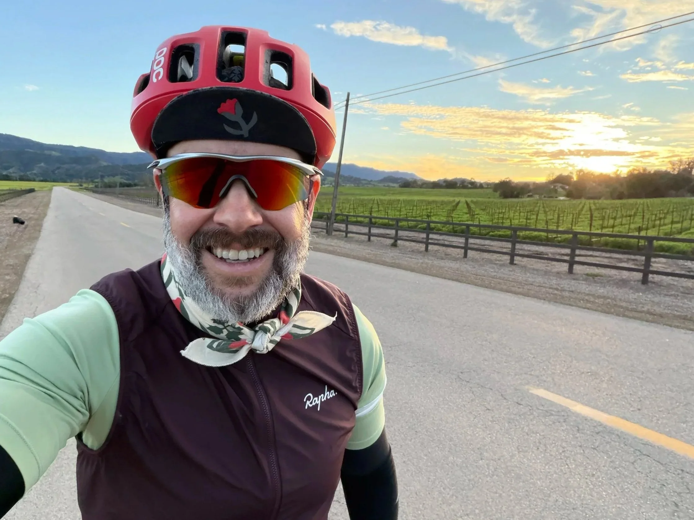 A smiling man wearing sunglasses, a red bike helmet, a light green and dark maroon cycling jersey, and a neck gaiter stands on an empty road with vineyards and hills in the background during sunset.
