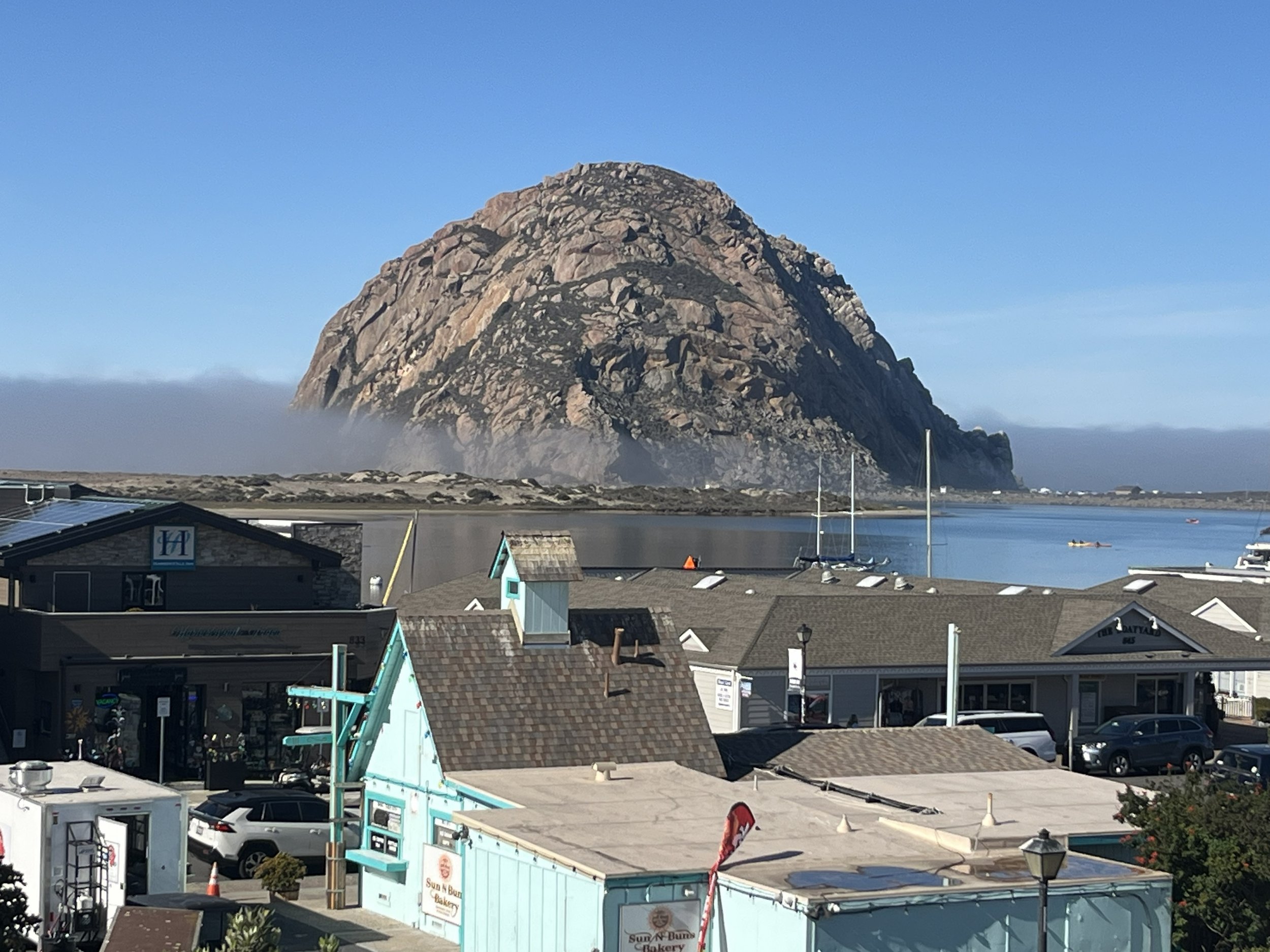 A large rocky mountain with a mostly clear blue sky in the background, and some clouds near the horizon. At the base of the mountain, there are buildings, boats on the water, and a parking lot with cars and a few boats docked nearby.