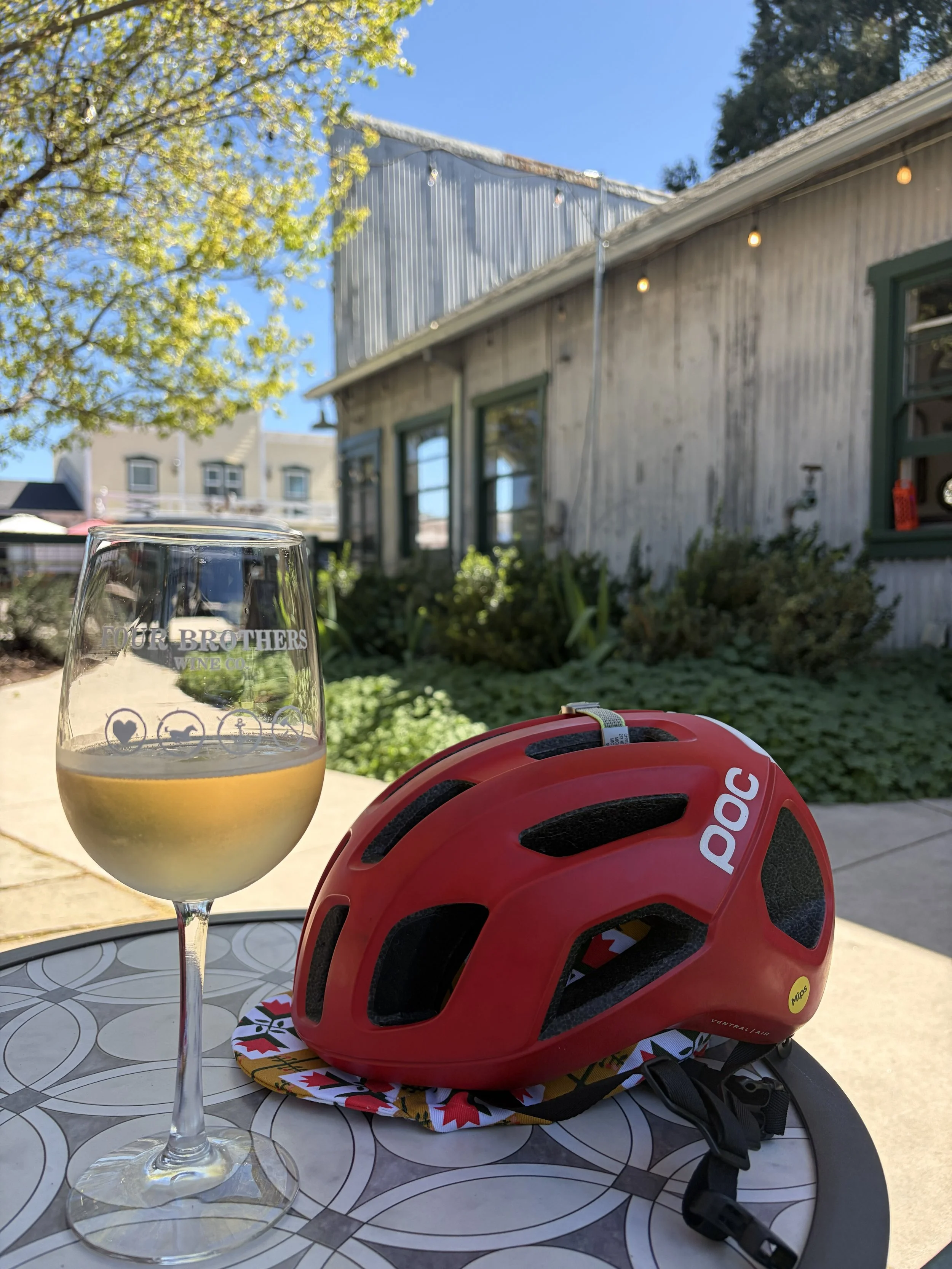 A glass of white wine, a red bicycle helmet, and a colorful cycling cap on a patterned table outdoors near a rustic building with string lights and trees.
