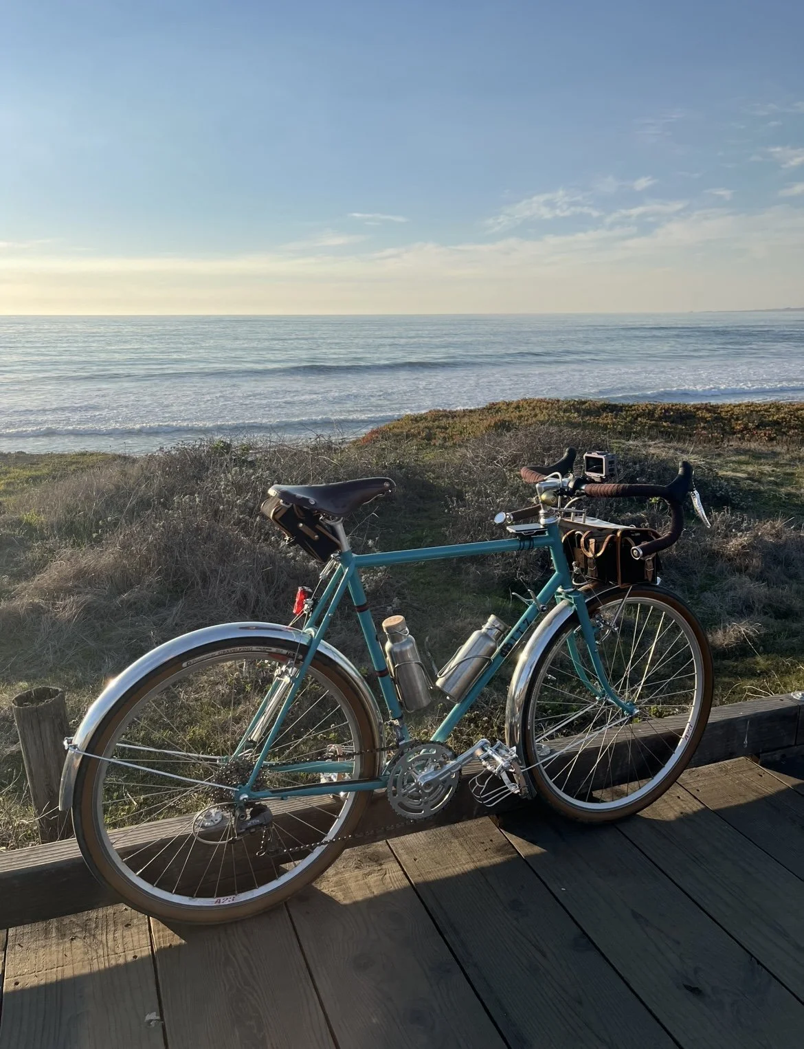 A teal bicycle resting on a wooden pathway near a coastal landscape with the ocean, hills, and a clear sky.