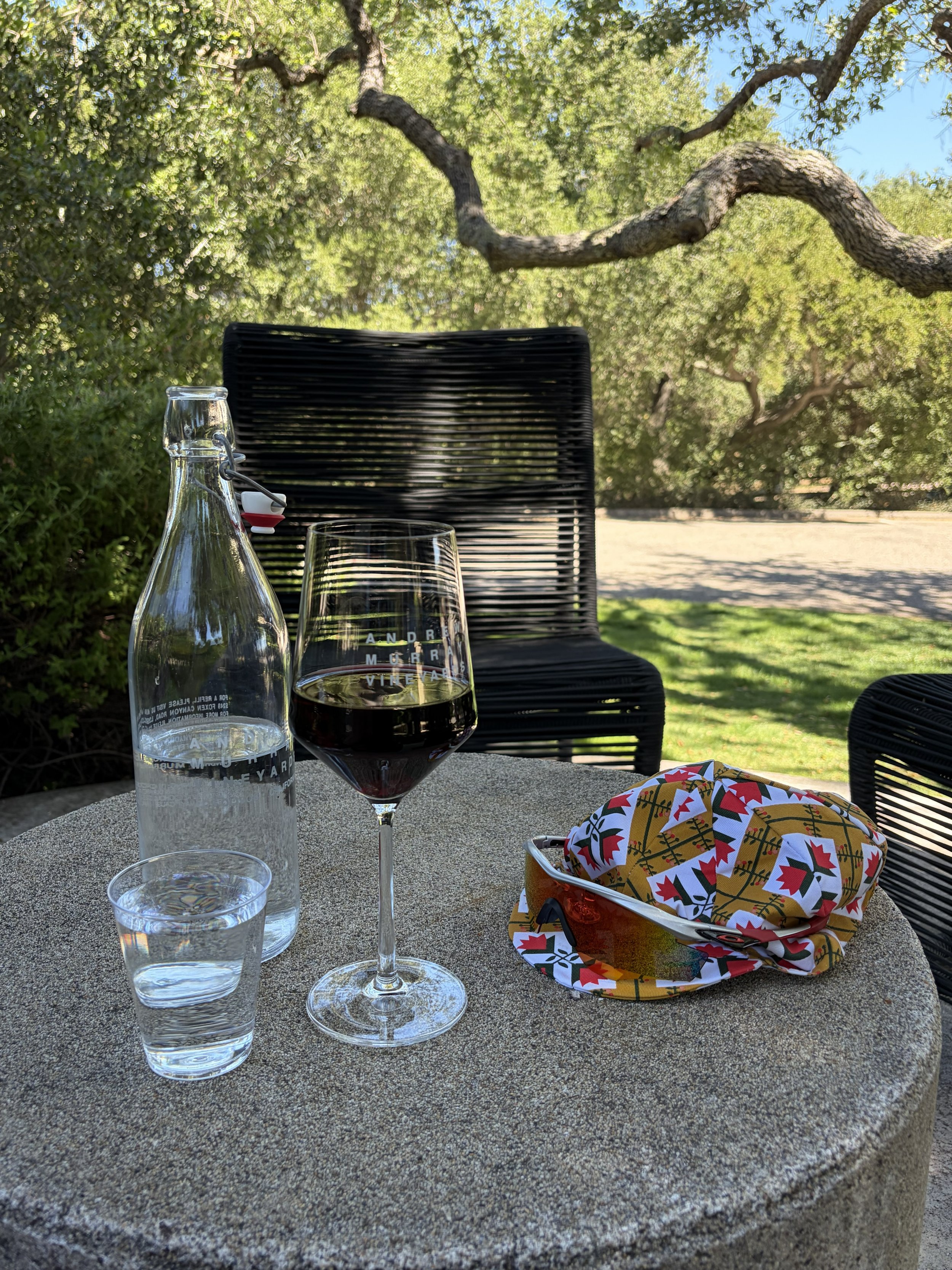 A table set outdoors with a glass of red wine, a water carafe, and a glass of water on a gravel surface. In the background, there is a black outdoor chair, a tree with green leaves, and a blue sky. A colorful hat with a festive pattern is on the tabl