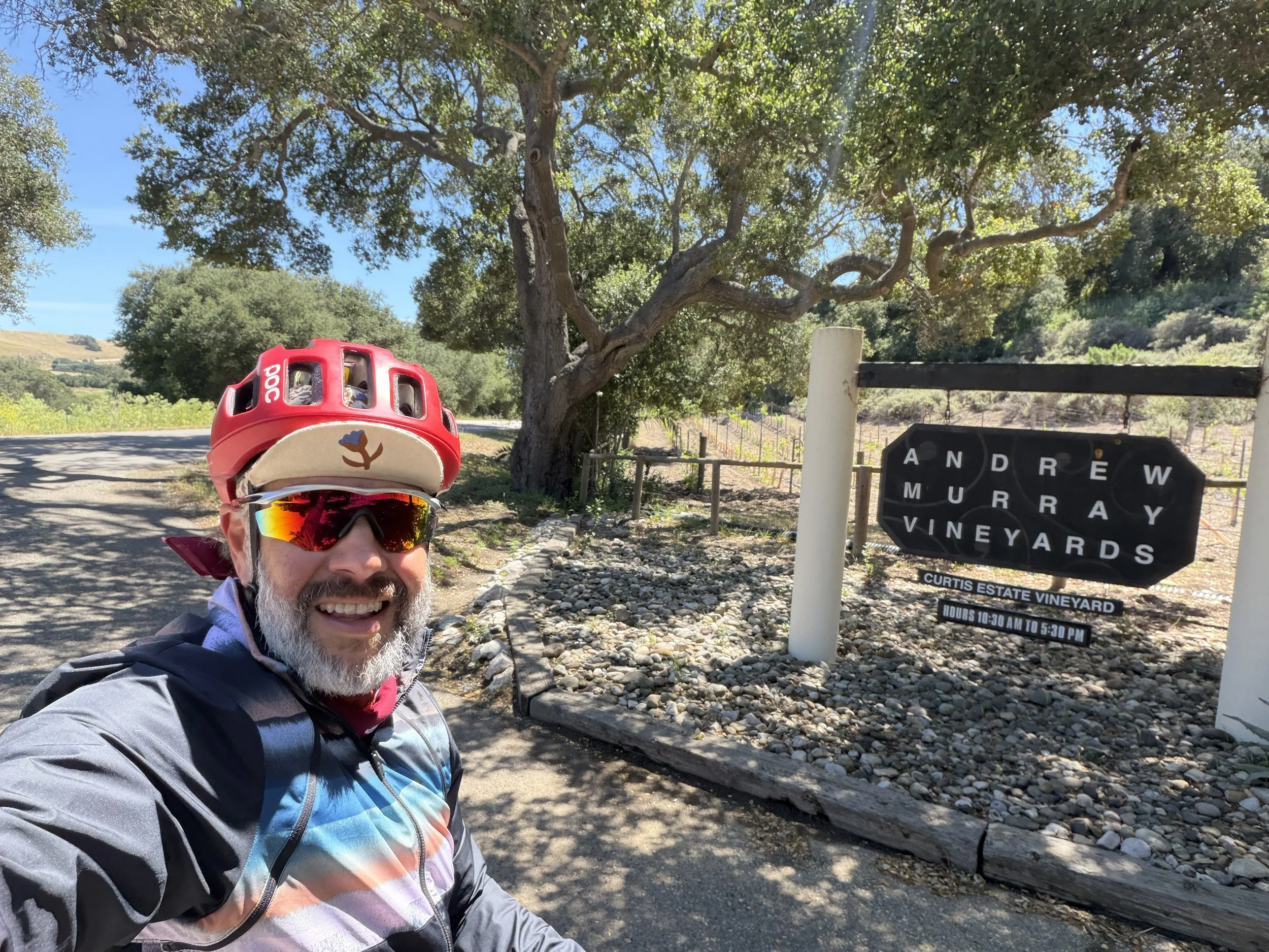 A smiling man with a beard taking a selfie outdoors. He is wearing a helmet with a logo, sunglasses, and a jacket. In the background, there are trees, a vineyard sign reading 'Andrew Murray Vineyards', and a clear blue sky.