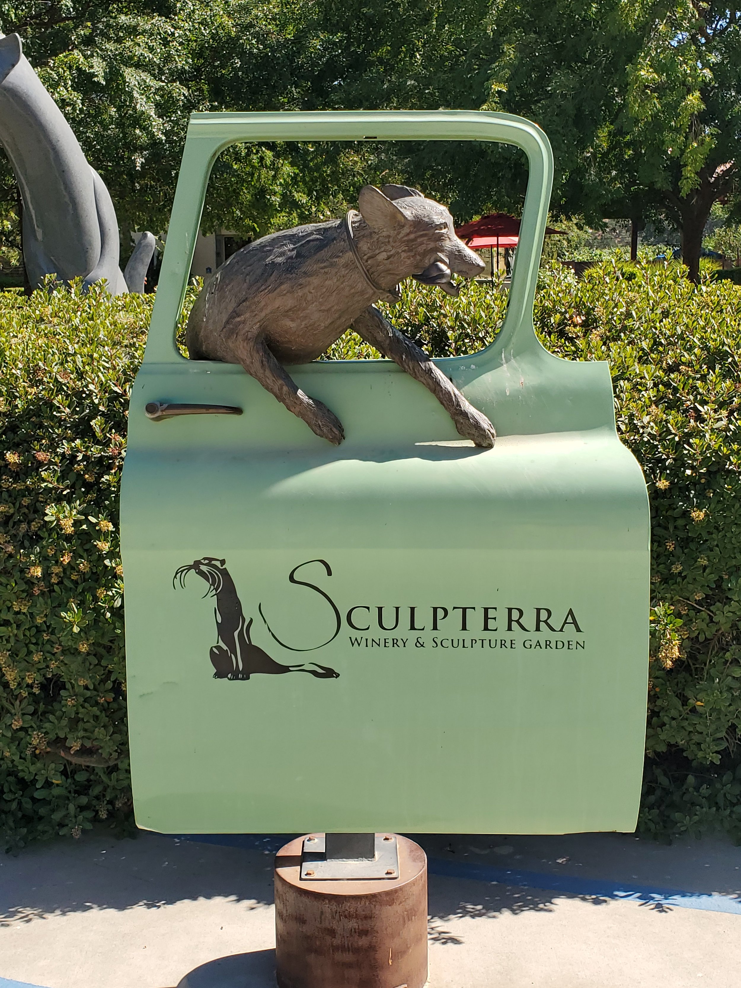 A sculpture of a wolf's head and front legs resting on top of a green sign that reads 'Sculptterra Winery & Sculpture Garden' with a black logo of a standing animal, possibly a wolf or dog, on the left side of the text. The sculpture appears to be ma