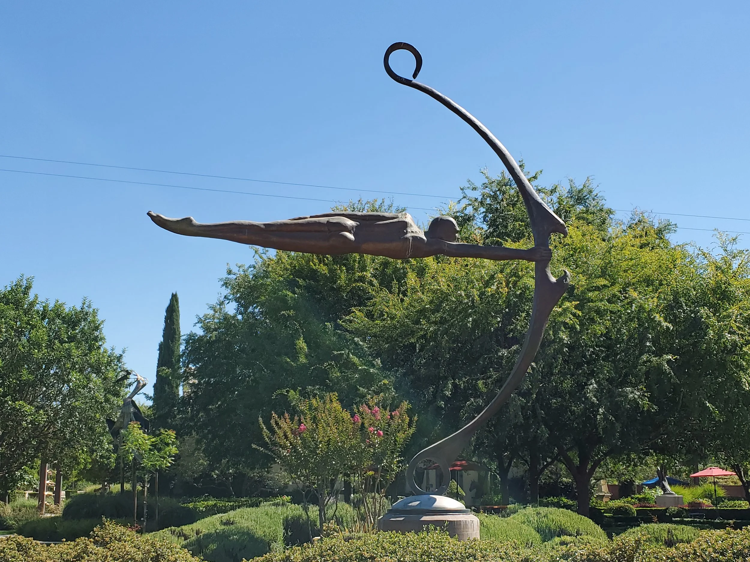 A metal sculpture of an abstract figure with elongated limbs and a circular element near the head, set among trees and greenery outdoors.