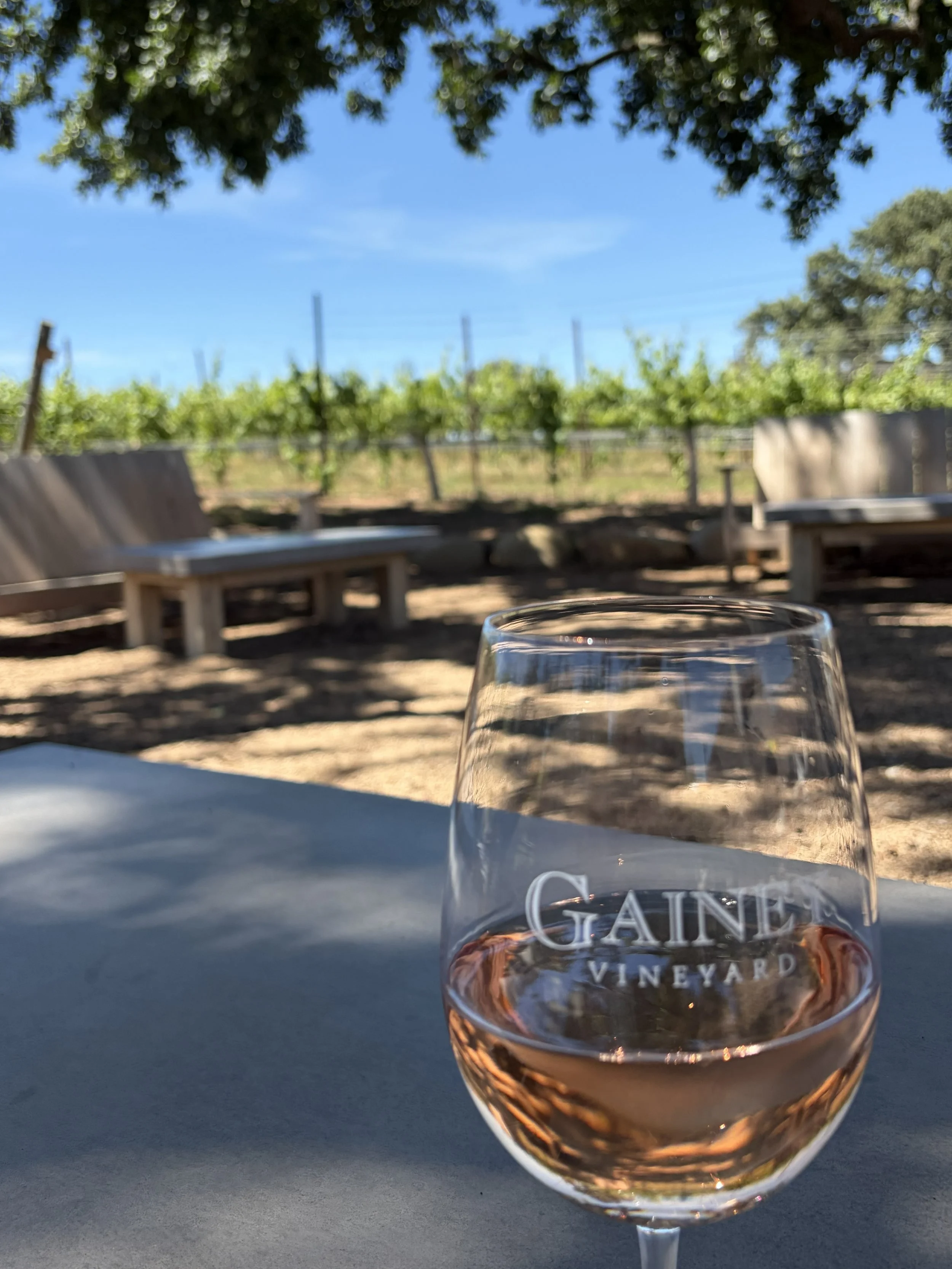 A glass of rosé wine on a table outdoors with a vineyard, greenery, and a blue sky in the background.