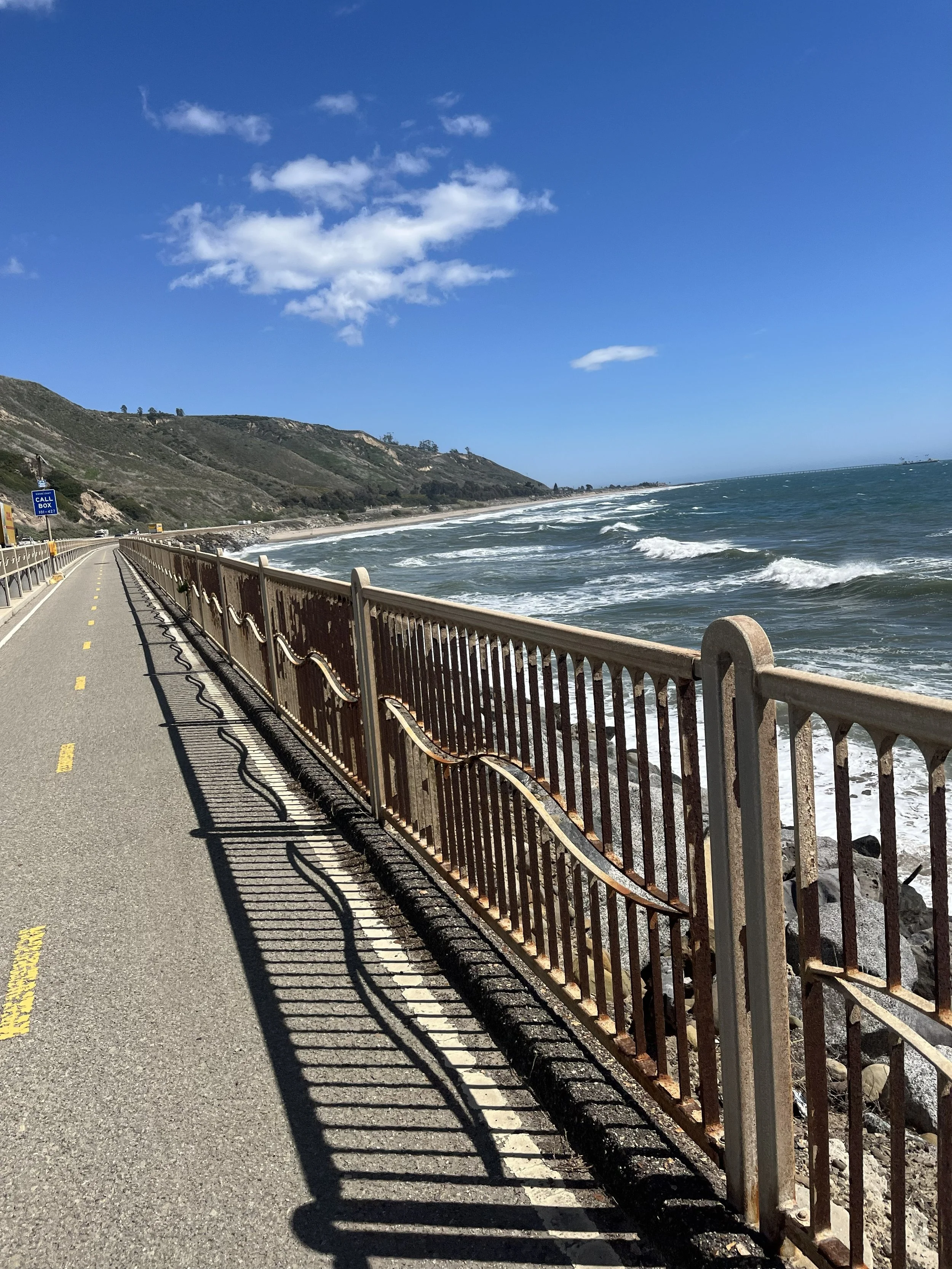 A coastal road with a yellow dividing line, a rusty metal railing, and ocean waves on the rocky shoreline under a clear blue sky with scattered clouds.