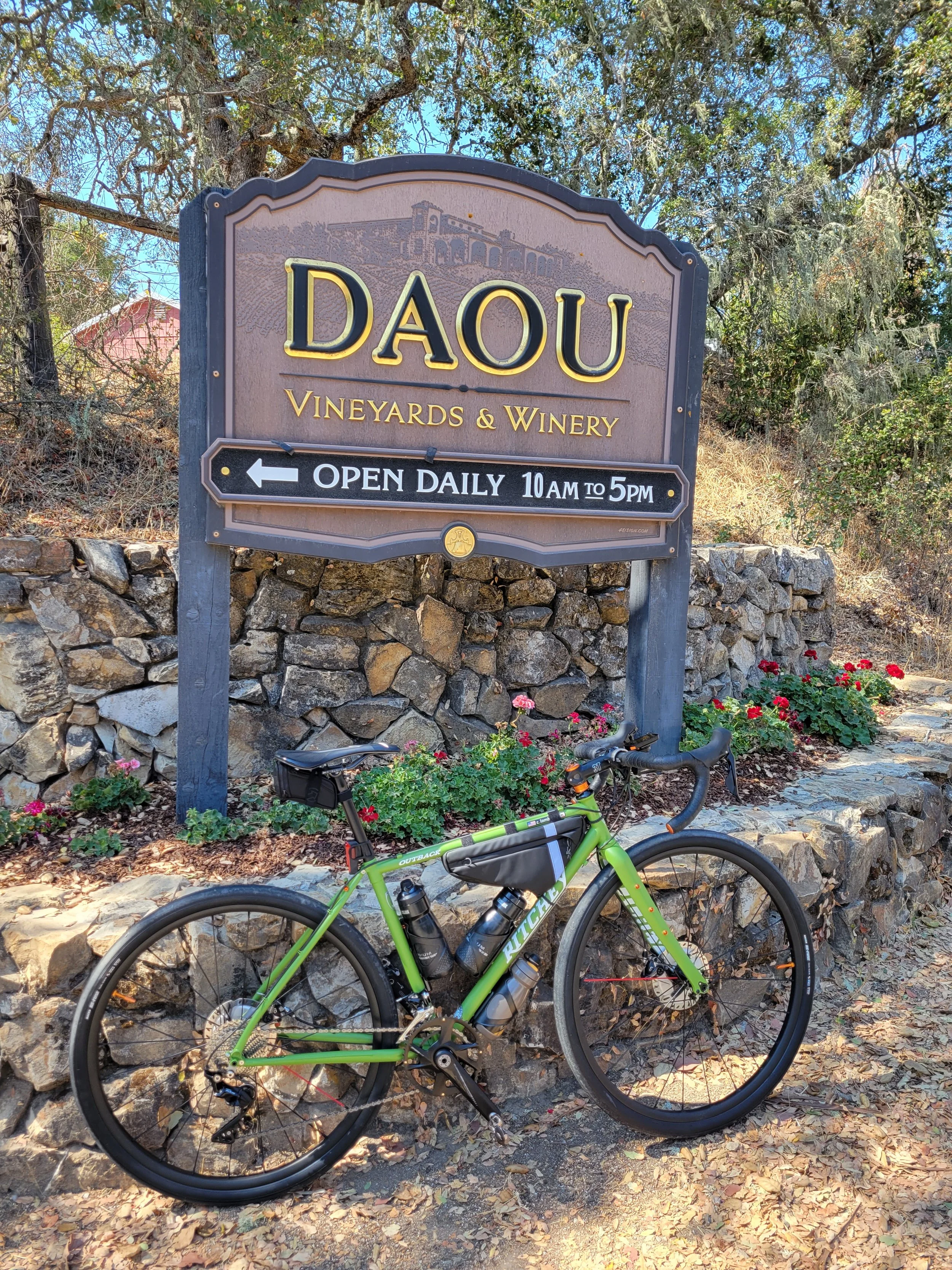 A green bicycle parked in front of a sign for Daou Vineyards & Winery, with a stone wall and trees in the background.