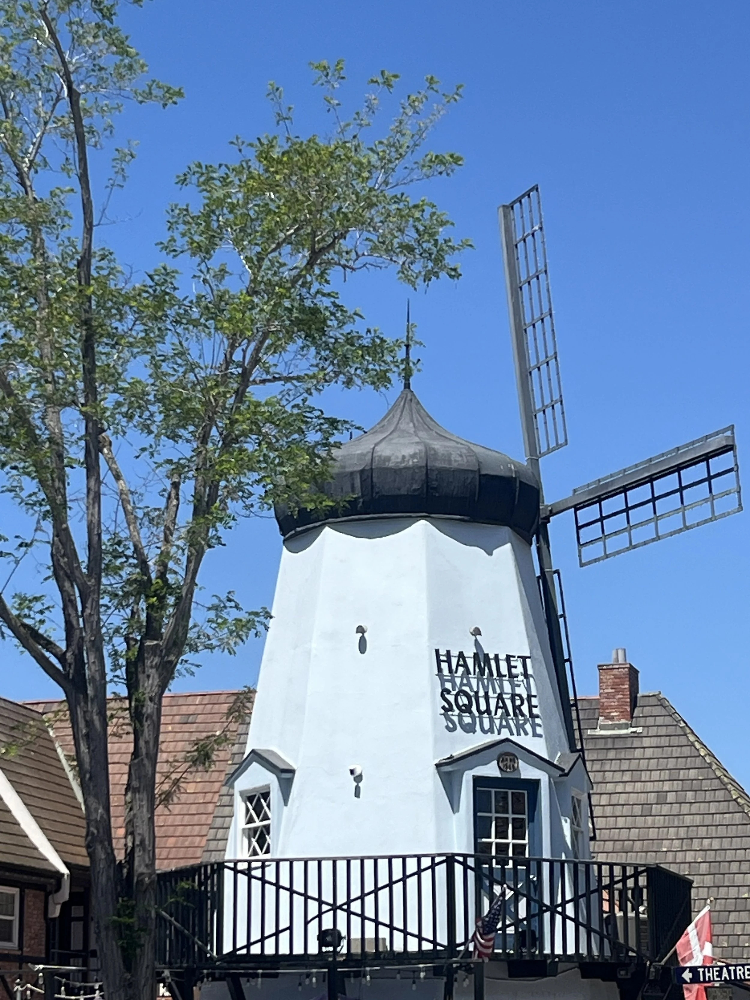 The image shows the Hamnet Shakespeare Cultural Center building with a prominent white tower, black railing, and signage that reads "HAMLET SQUARE". There are trees and rooftops of nearby buildings as part of the scene.