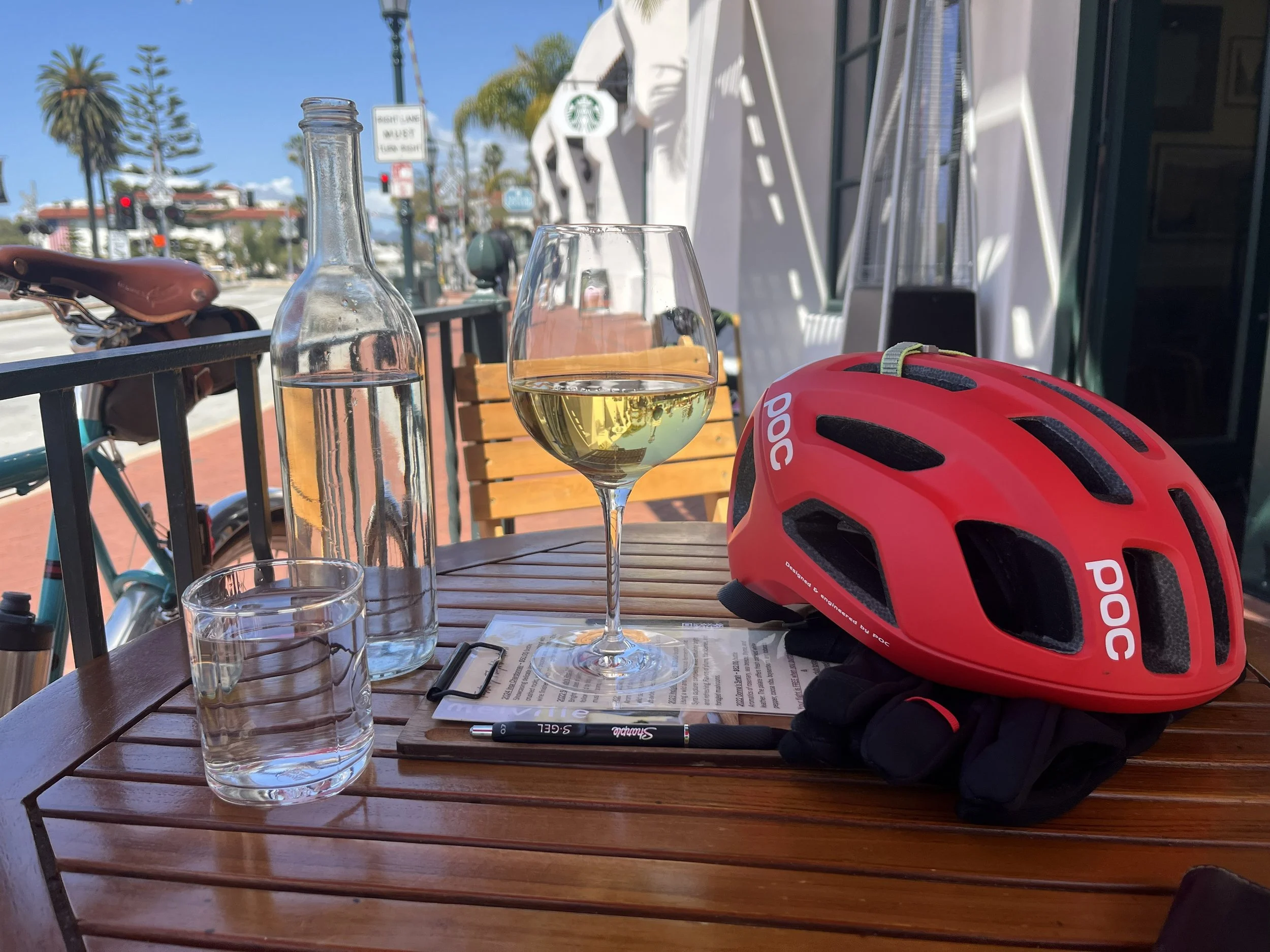 A wooden outdoor patio table with a glass of white wine, a bottle of water, a red bicycle helmet, a black marker, and some papers. In the background, there are white umbrellas, palm trees, and a street scene with traffic lights and buildings under a 