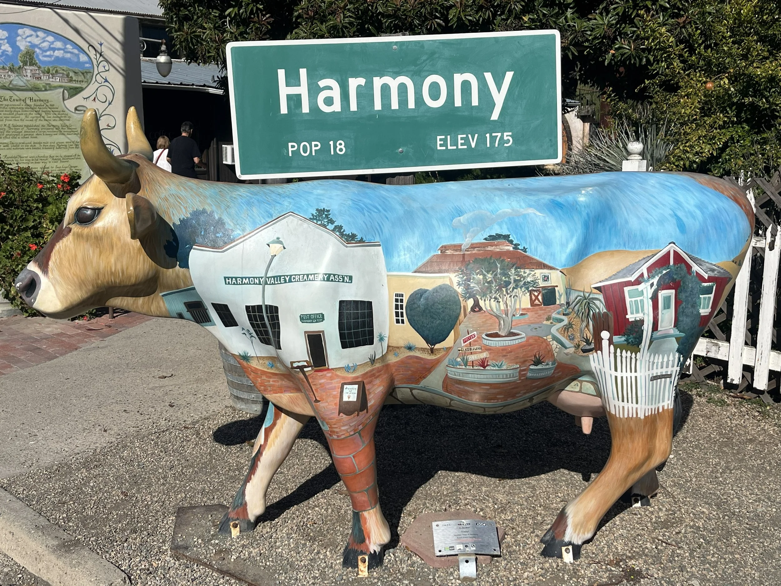 A decorative cow sculpture painted with a mural of houses, trees, clouds, and a blue sky, on a street corner in Harmony, California.