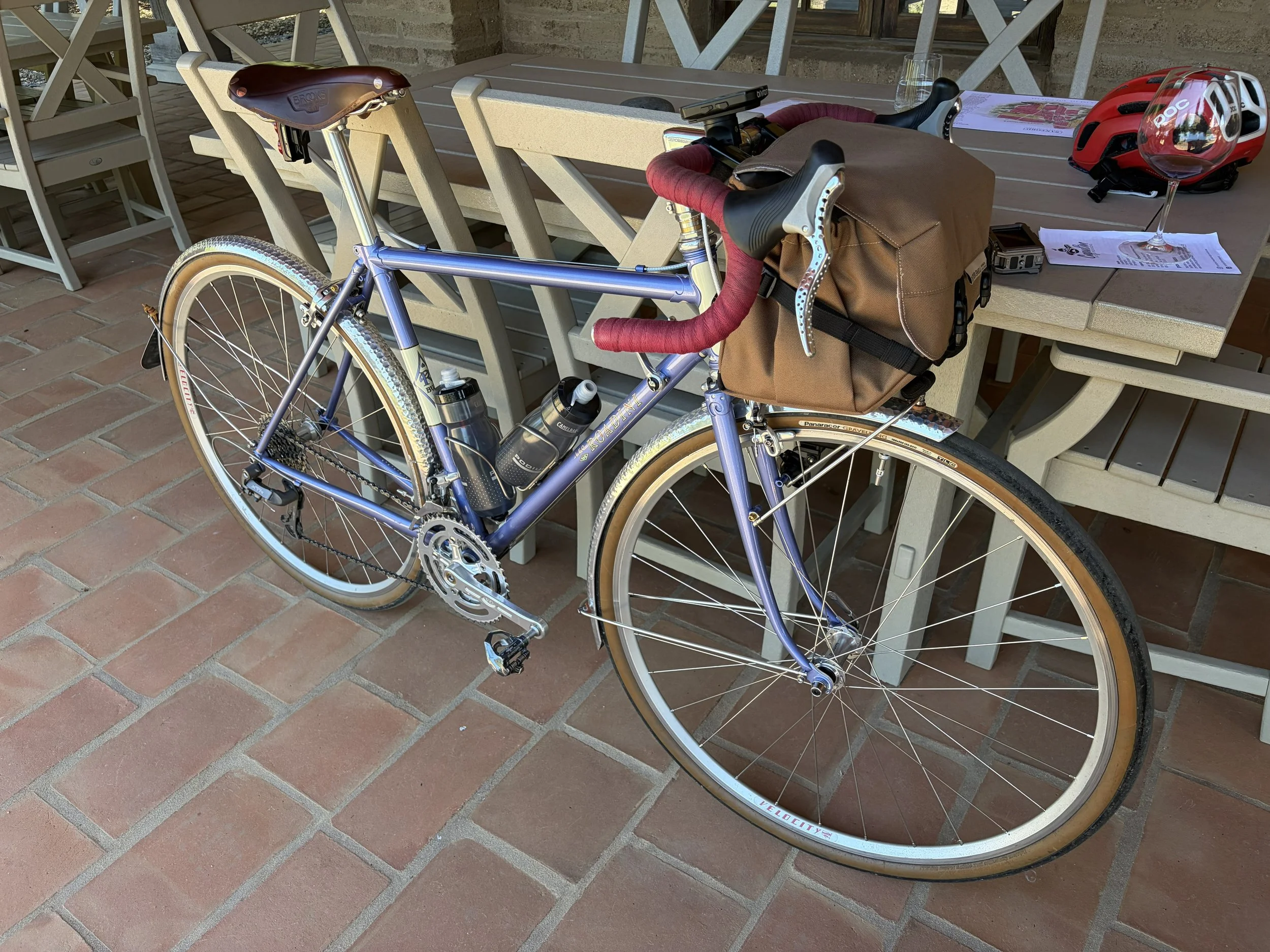 A blue and silver road bicycle is parked against a beige outdoor dining table on a brick patio. The bike has a brown saddle and a brown handlebar bag, with a red helmet, a wine glass, and some papers and a camera on the table nearby.