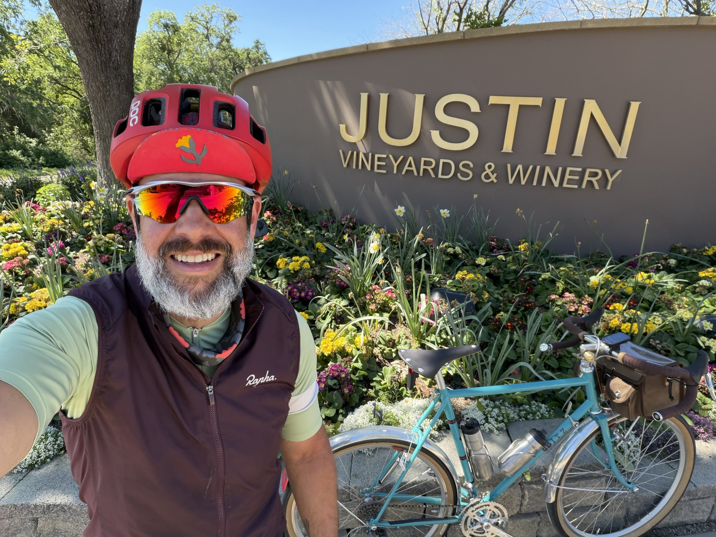 A man with a beard taking a selfie in front of a sign that reads "Justin Vineyards & Winery." He is wearing a red cycling helmet, sunglasses, and a black vest over a light green shirt. There are colorful flowers and a blue bicycle in the background, 
