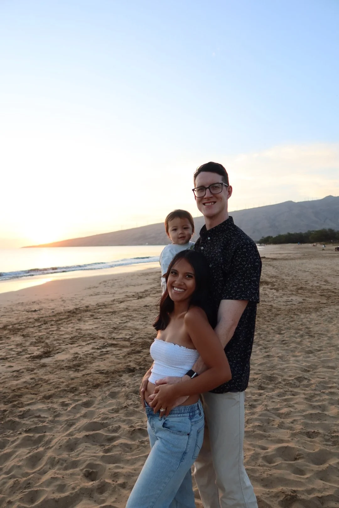 A smiling family of three standing on a sandy beach during sunset. The woman is in front, wearing a white strapless top and jeans, with one hand on her stomach. The man is behind her, wearing a dark patterned shirt and khakis, holding a young child with short hair and a white top. The background shows calm ocean waves, a mountain range, and a colorful sky at sunset.