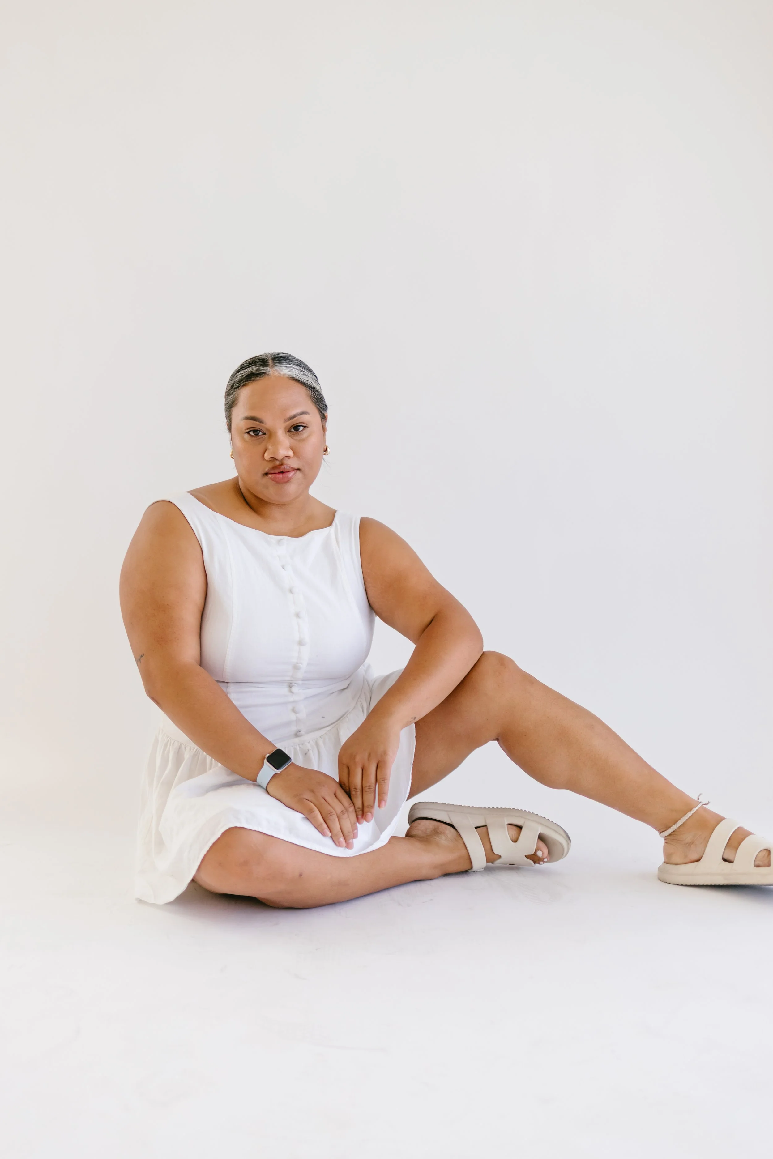 A woman wearing a white dress, sitting on the floor with one knee raised, against a plain white background.
