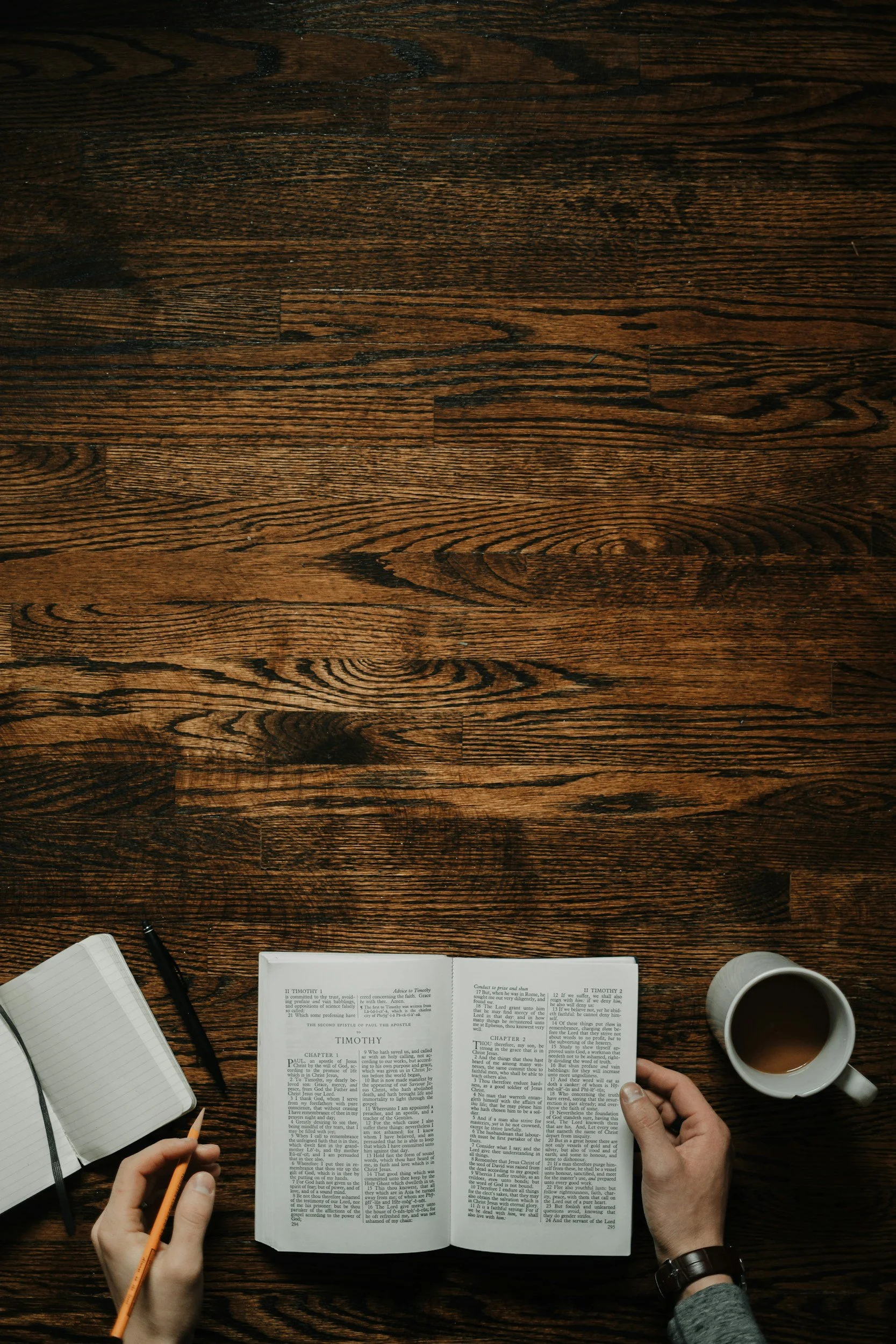 Overhead view of a wooden table with open books, a person's hands holding a pencil and a coffee mug, and a notepad.