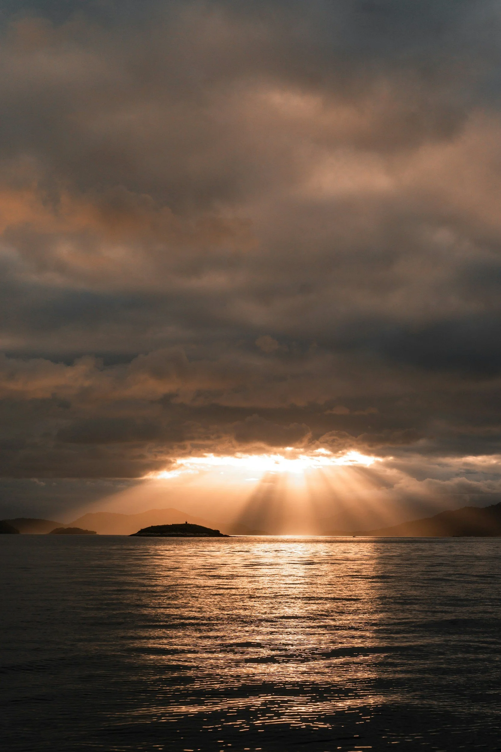 Sunset over the ocean with rays of sunlight breaking through dark, cloudy skies and distant islands on the horizon.