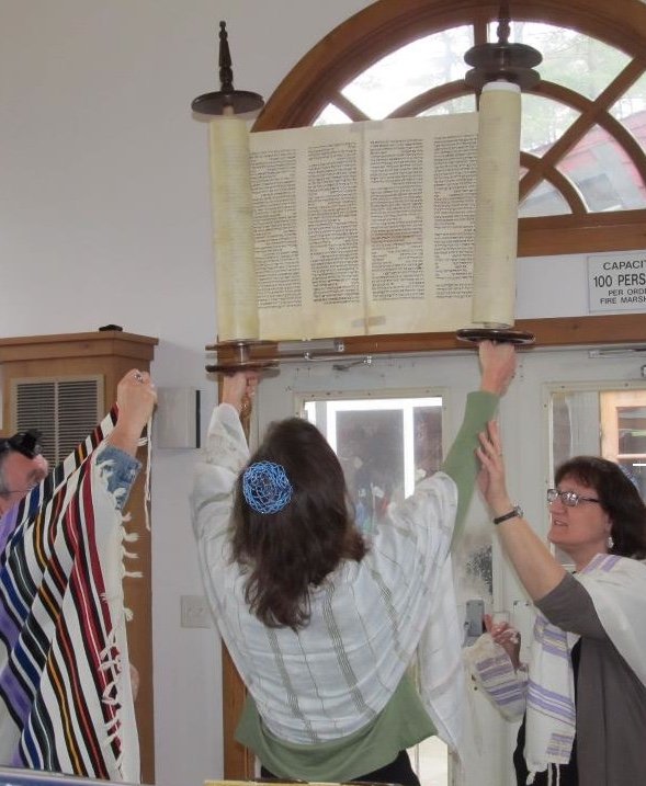 Three women are holding up a large Torah scroll inside a building with a wooden arch window. One woman is wearing a green and white shawl, another is in a striped shawl, and the third is in glasses and a scarf. The Torah is open with scriptural text, and sunlight is coming through the window.