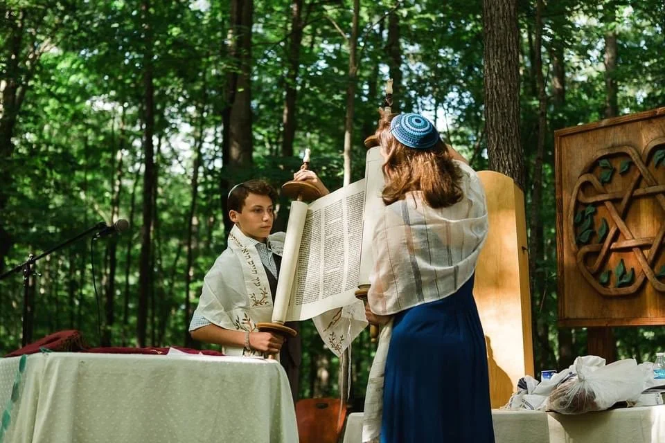 A boy and a woman in a wooded outdoor setting during a religious ceremony, with the woman holding a Torah scroll.