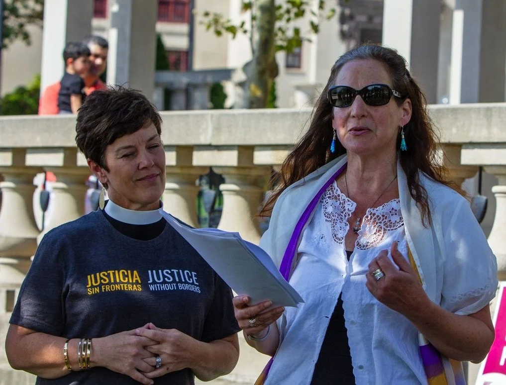 Two women speaking outdoors, one wearing a black shirt with 'Justice Without Borders' written in English and Spanish, and the other dressed in a white blouse and sunglasses, holding papers, with a man and a boy in the background.