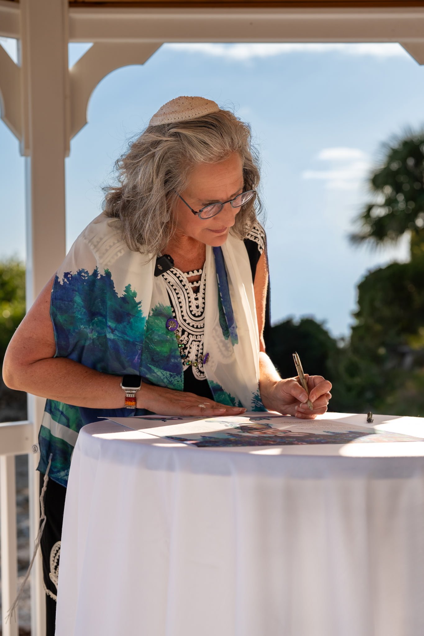 An older woman with gray hair, glasses, and wearing a patterned dress and a white shawl with a scenic print, is signing a document on a white table outdoors under a porch with a blue sky and greenery in the background.