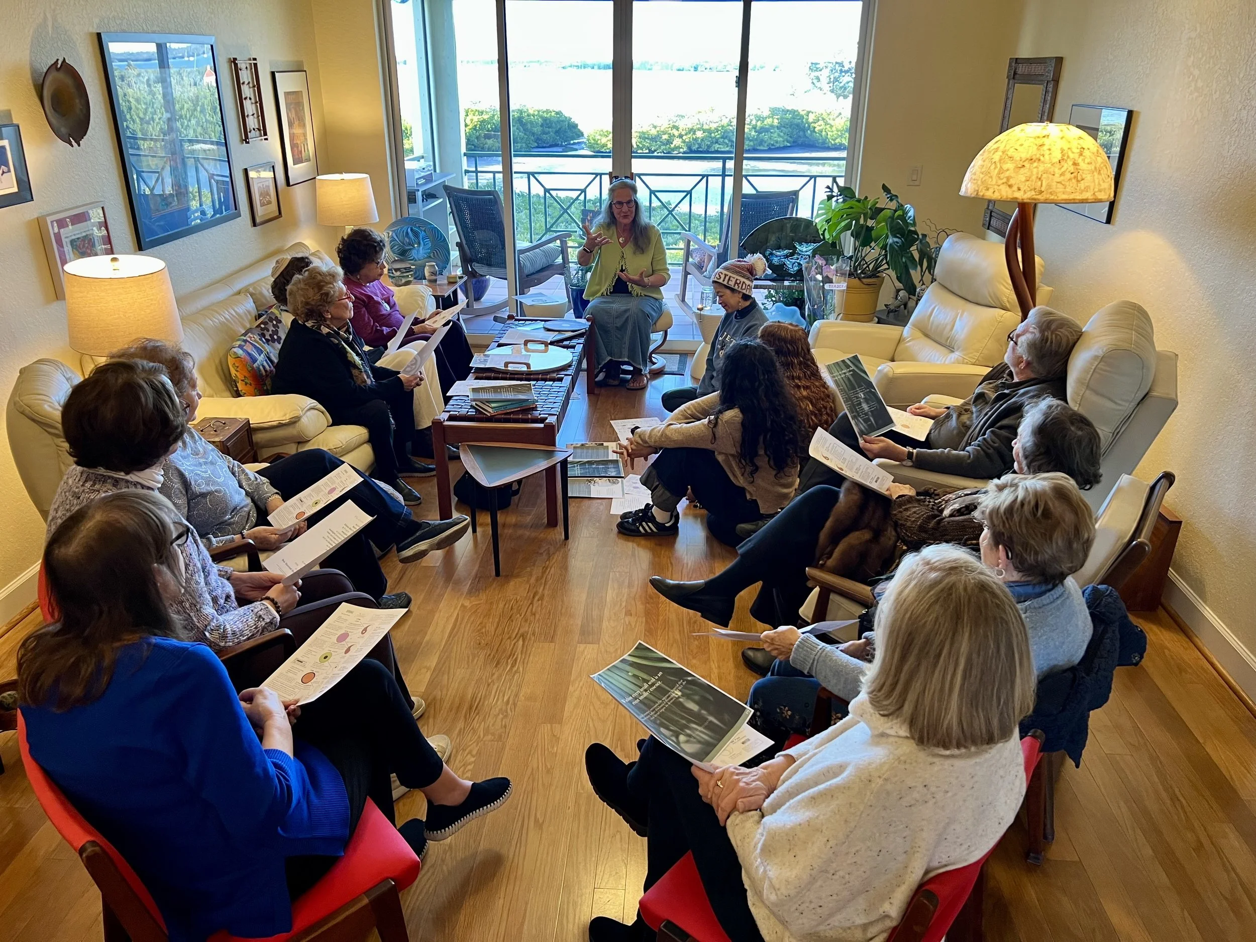 A group of people sitting in a living room, participating in a discussion or lesson, with a woman standing in front of them speaking. The room is bright with a large window and balcony, decorated with wall art, lamps, and plants.