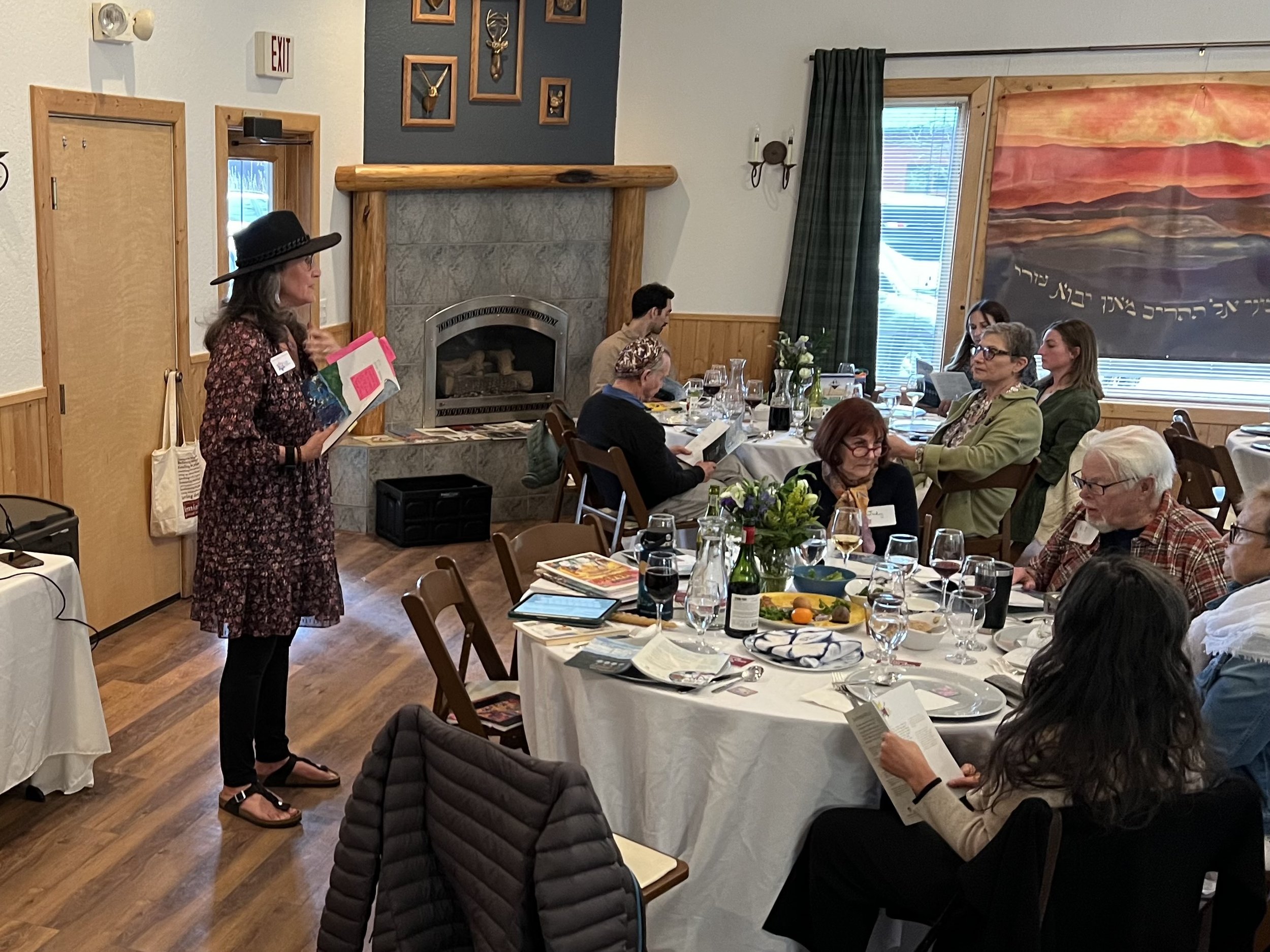 A woman in a floral dress and black hat stands in front of seated people at a table, speaking to the group during a gathering or meal event in a decorated room with artwork, a fireplace, and a large colorful painting on the wall.