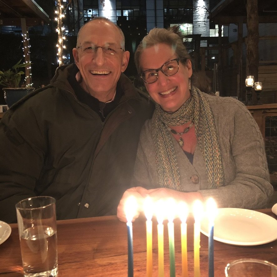 Two people smiling at a table with birthday candles, a glass of water, and plates in a restaurant with string lights in the background.