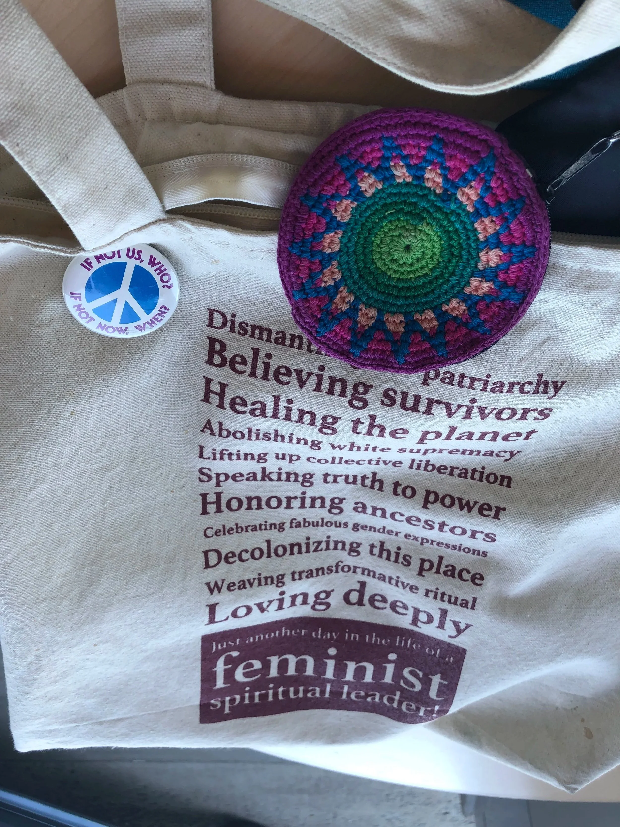 Close-up of a beige tote bag with a colorful crocheted pin on it. The tote bag has printed text about feminism, spirituality, and social issues.