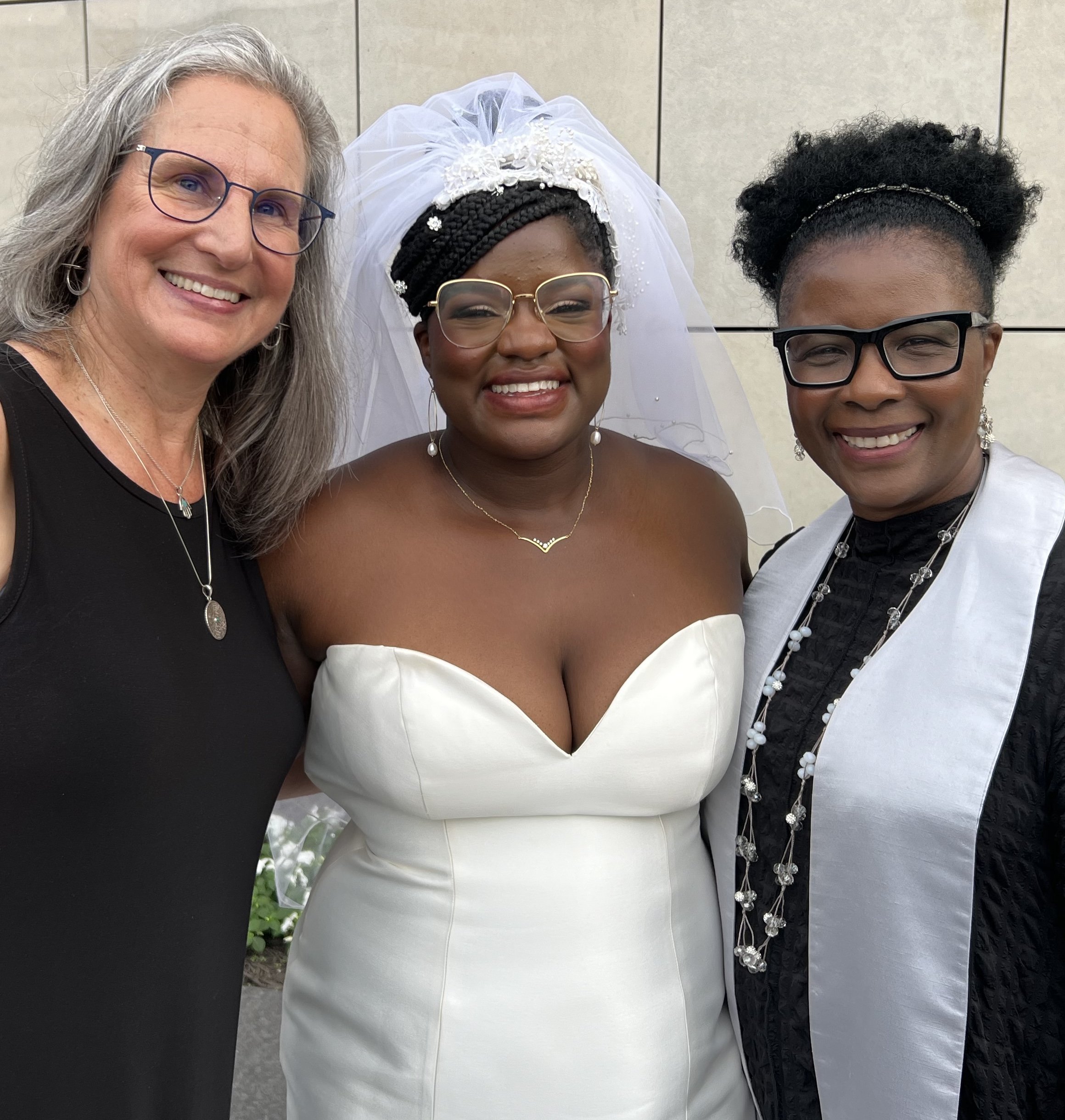 Three women smiling for a selfie, the woman in the middle is wearing a wedding dress and veil, and the women on either side are dressed in formal attire with jewelry.