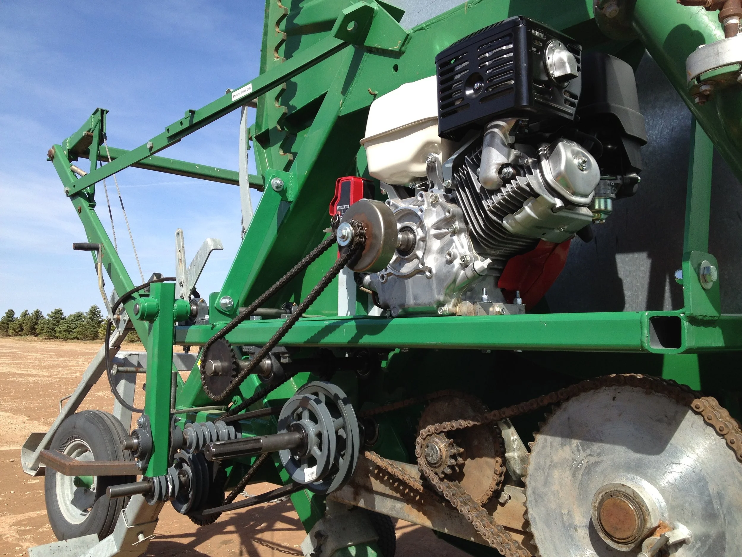Close-up of a green farm machinery frame with an exposed small engine, chains, and gears, against a background of an open field and blue sky.