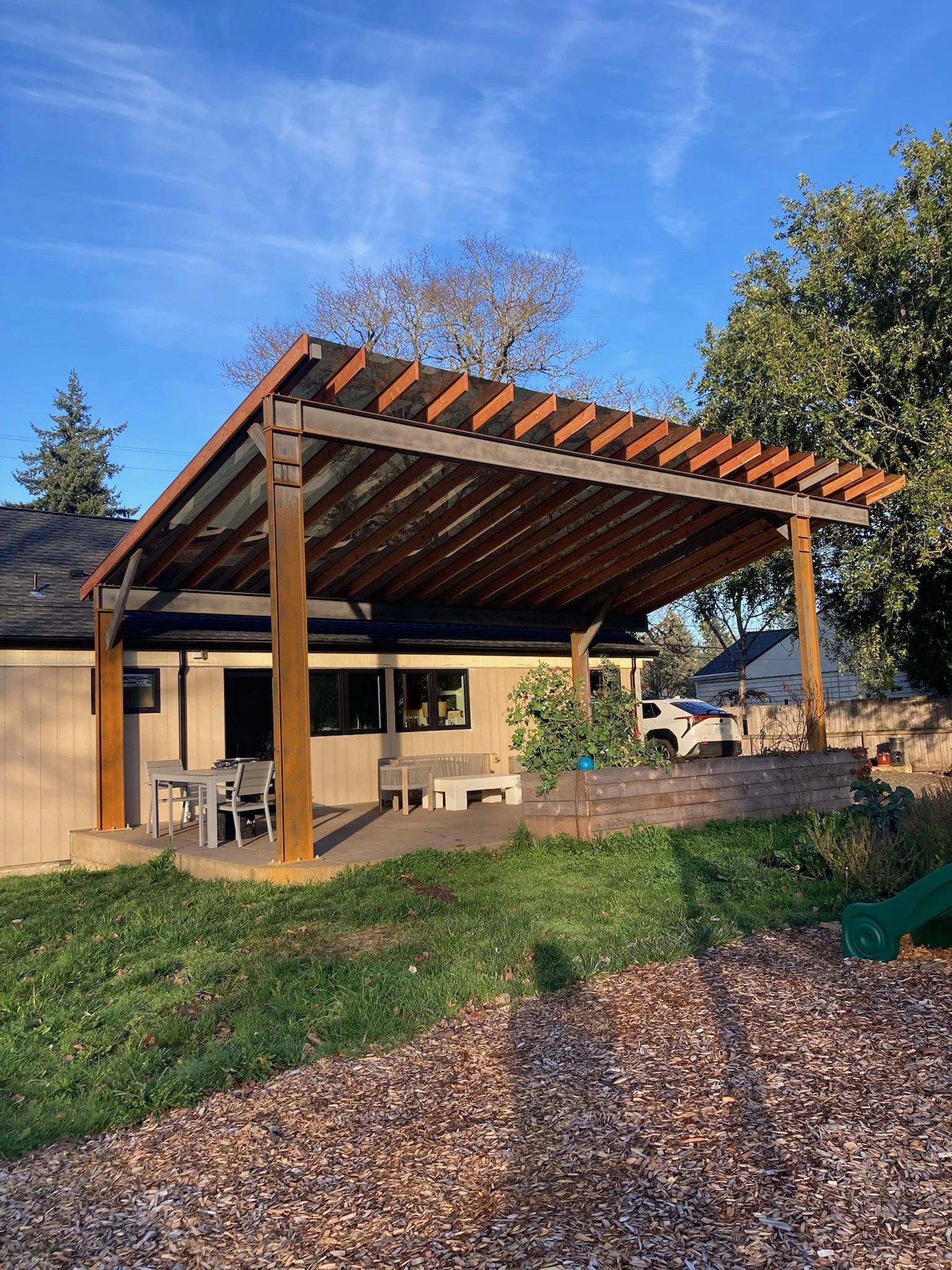 A backyard patio with a new wooden pergola, outdoor dining table and chairs, a raised garden bed with plants, and a white truck parked in the driveway under a clear blue sky.