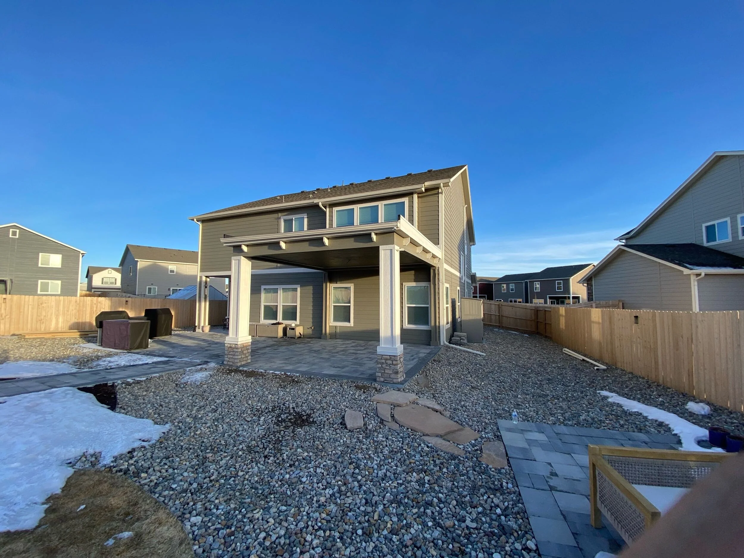 Backyard of a house with a stone patio, fencing, and several neighboring houses, during daytime with clear blue sky.