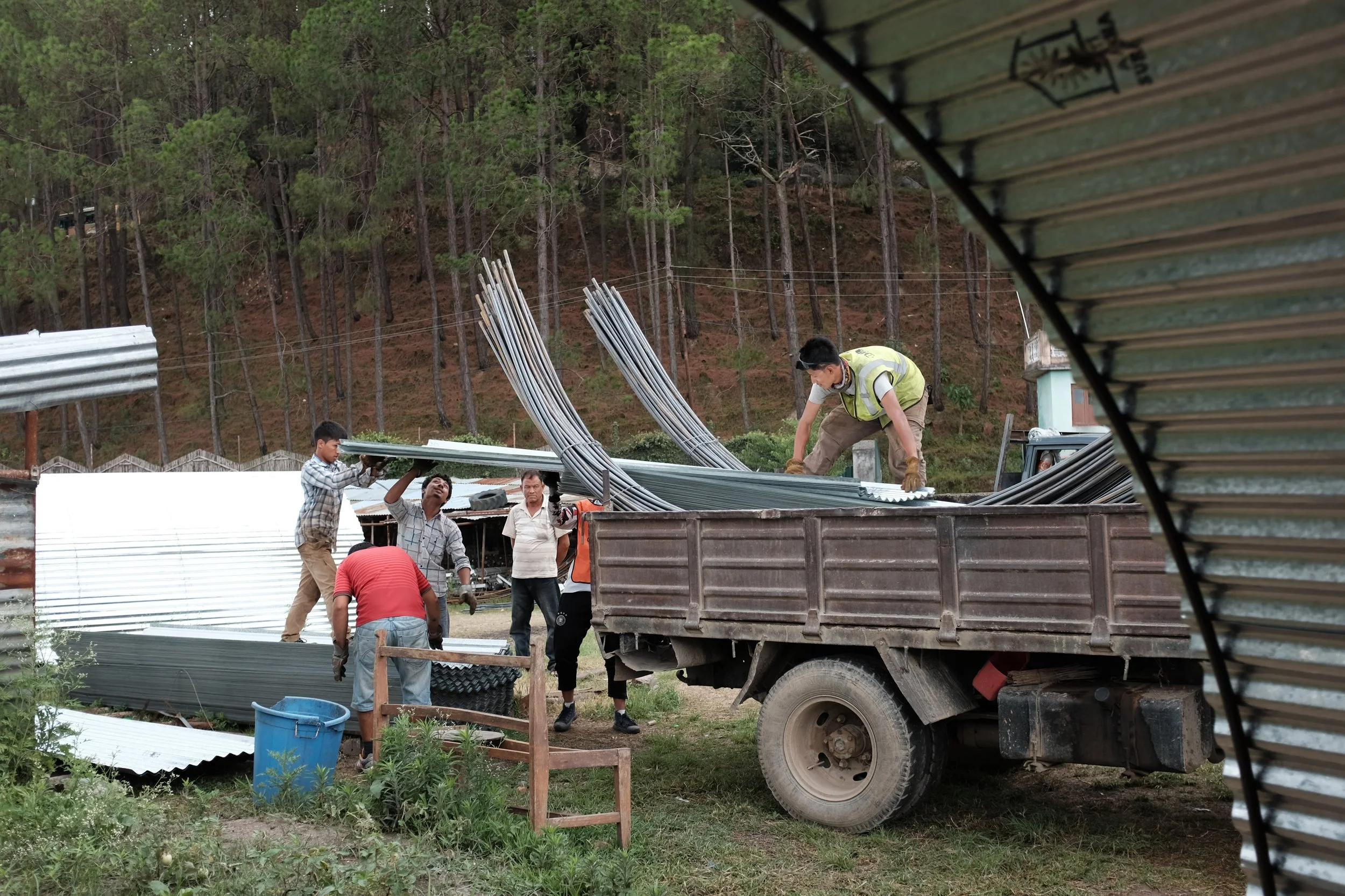 Several workers unload metal roofing sheets and long metal rods from a truck at a construction site in a rural area with trees in the background.
