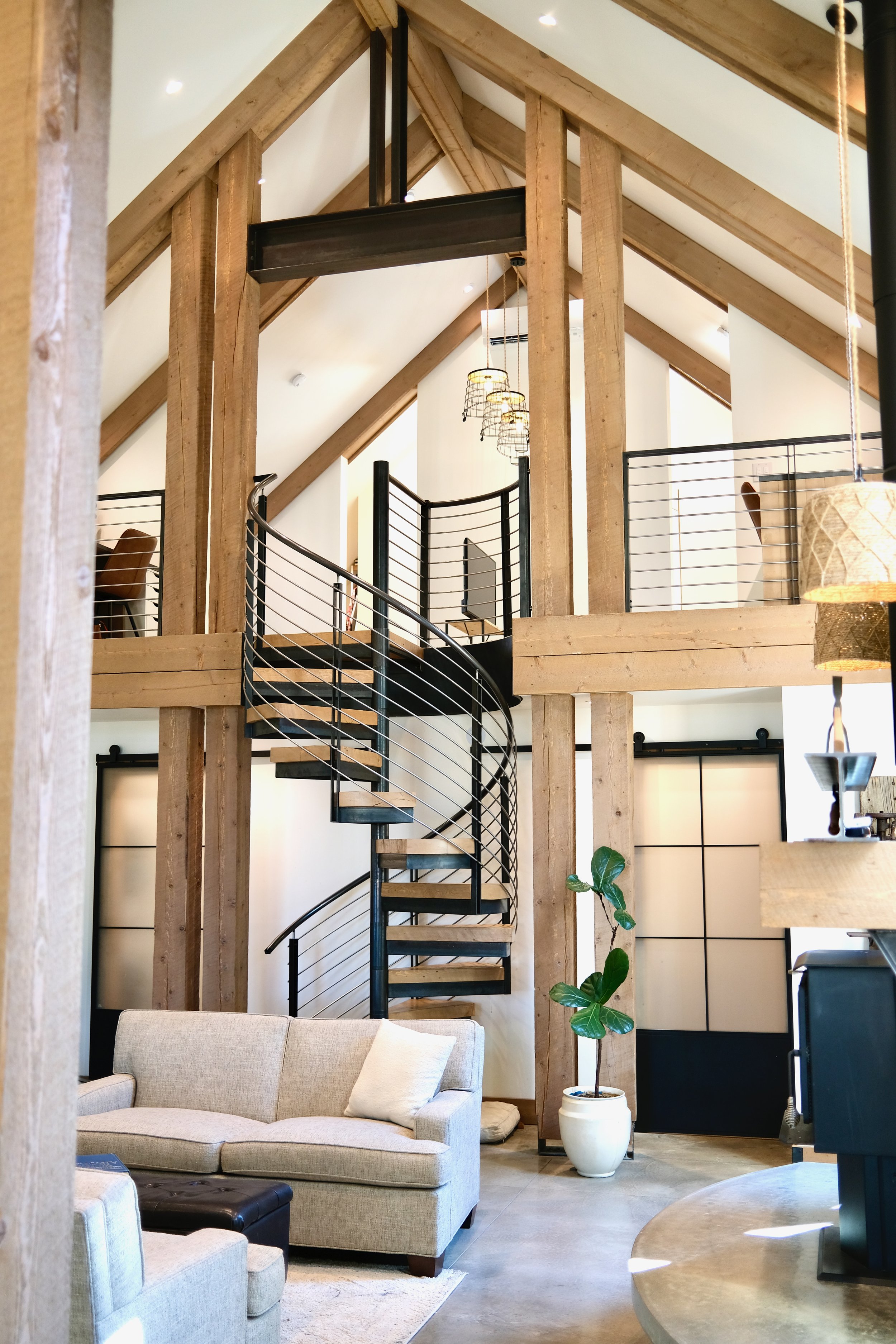 Interior of a modern home with a spiral staircase, exposed wood beams, a beige sofa, a potted plant, and decorative hanging lamps.